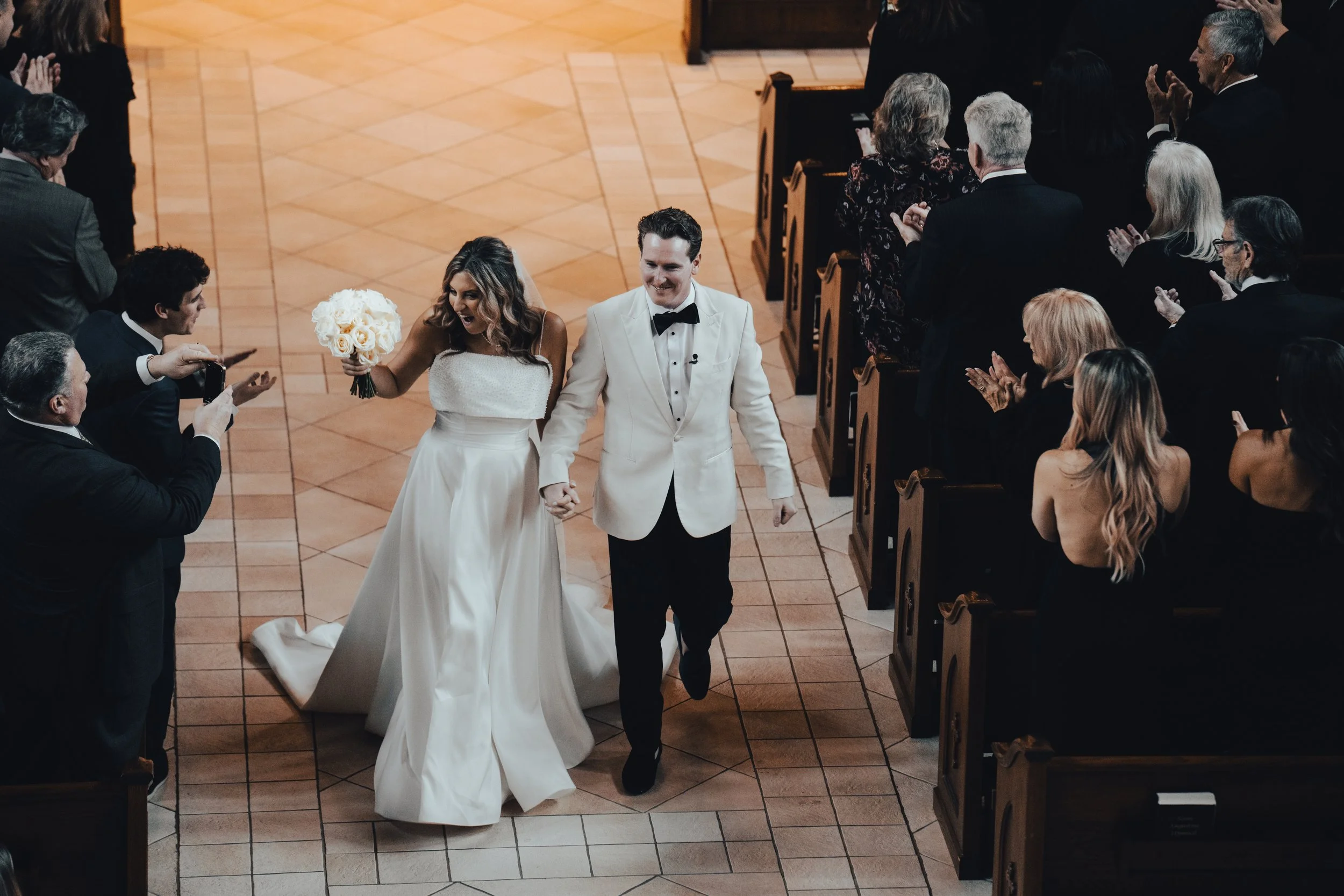 A newlywed couple walking down the aisle after their wedding ceremony, holding hands, with the bride holding a bouquet of white roses, surrounded by seated guests clapping and celebrating.