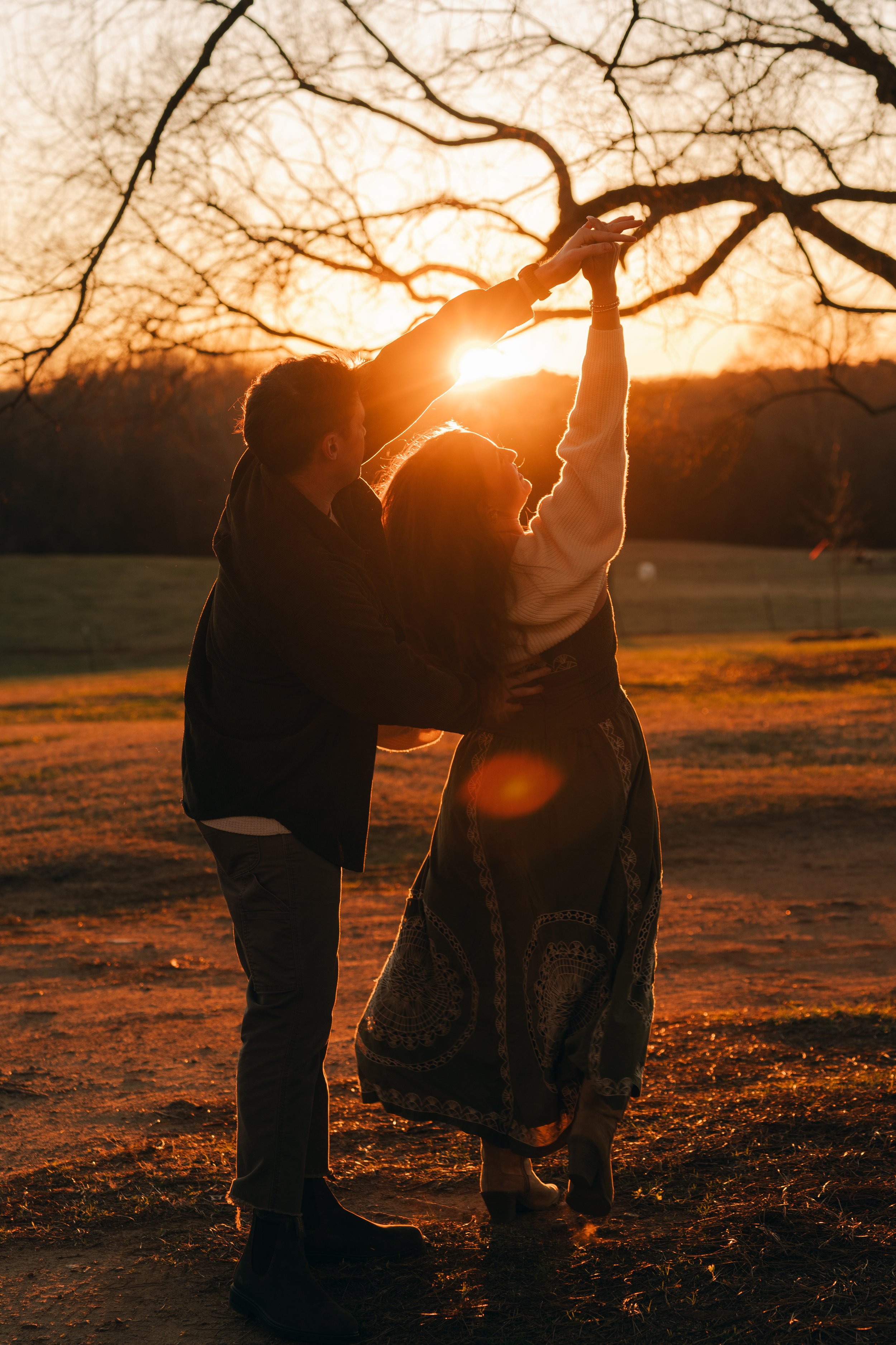Two people, a man and a woman, dancing outdoors during sunset with leafless trees in the background.