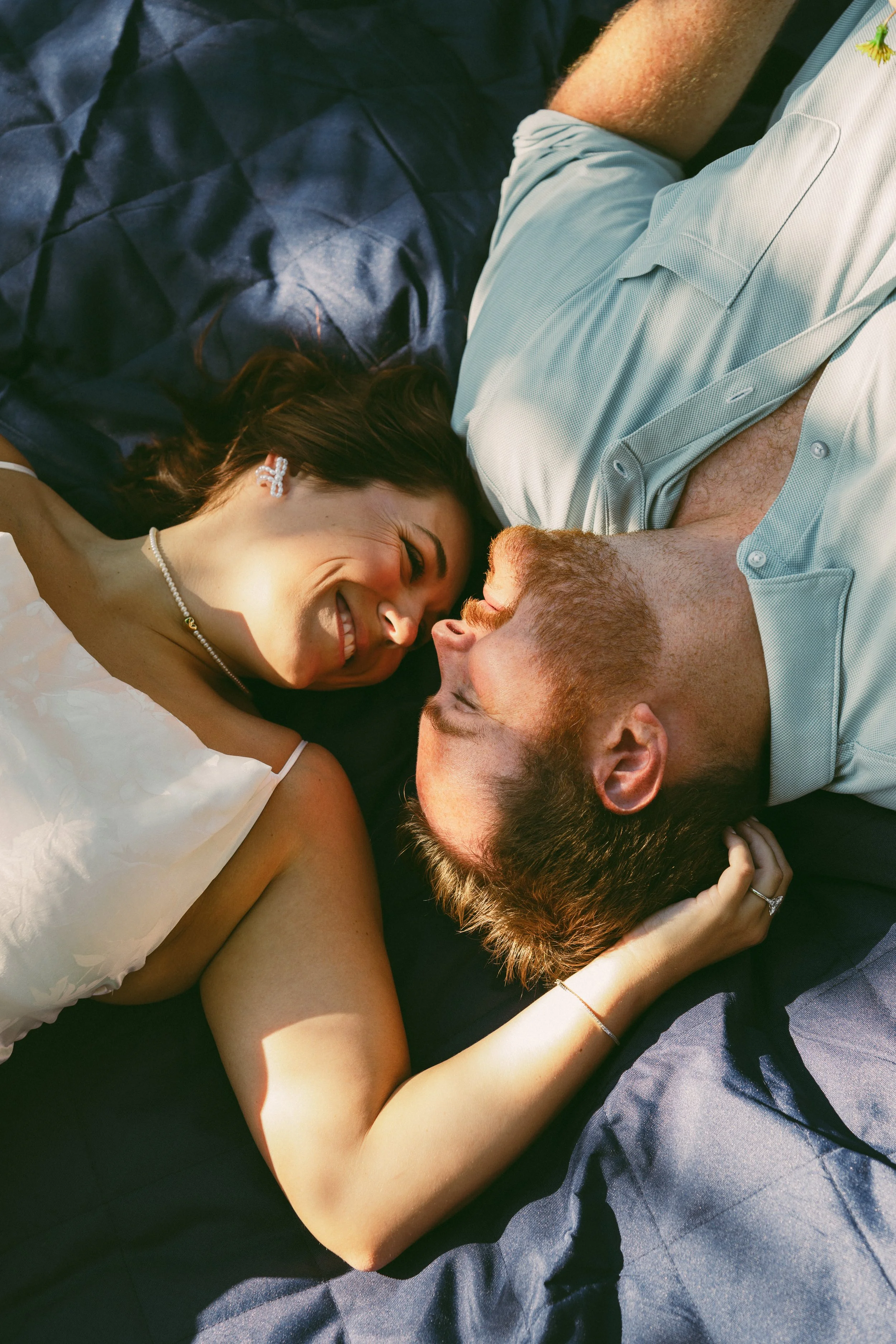 A smiling woman and a man are lying on their sides on a dark fabric surface, facing each other with their faces close together. The woman is wearing a white dress and pearl earrings, and the man has a beard and short hair. They are holding hands and 