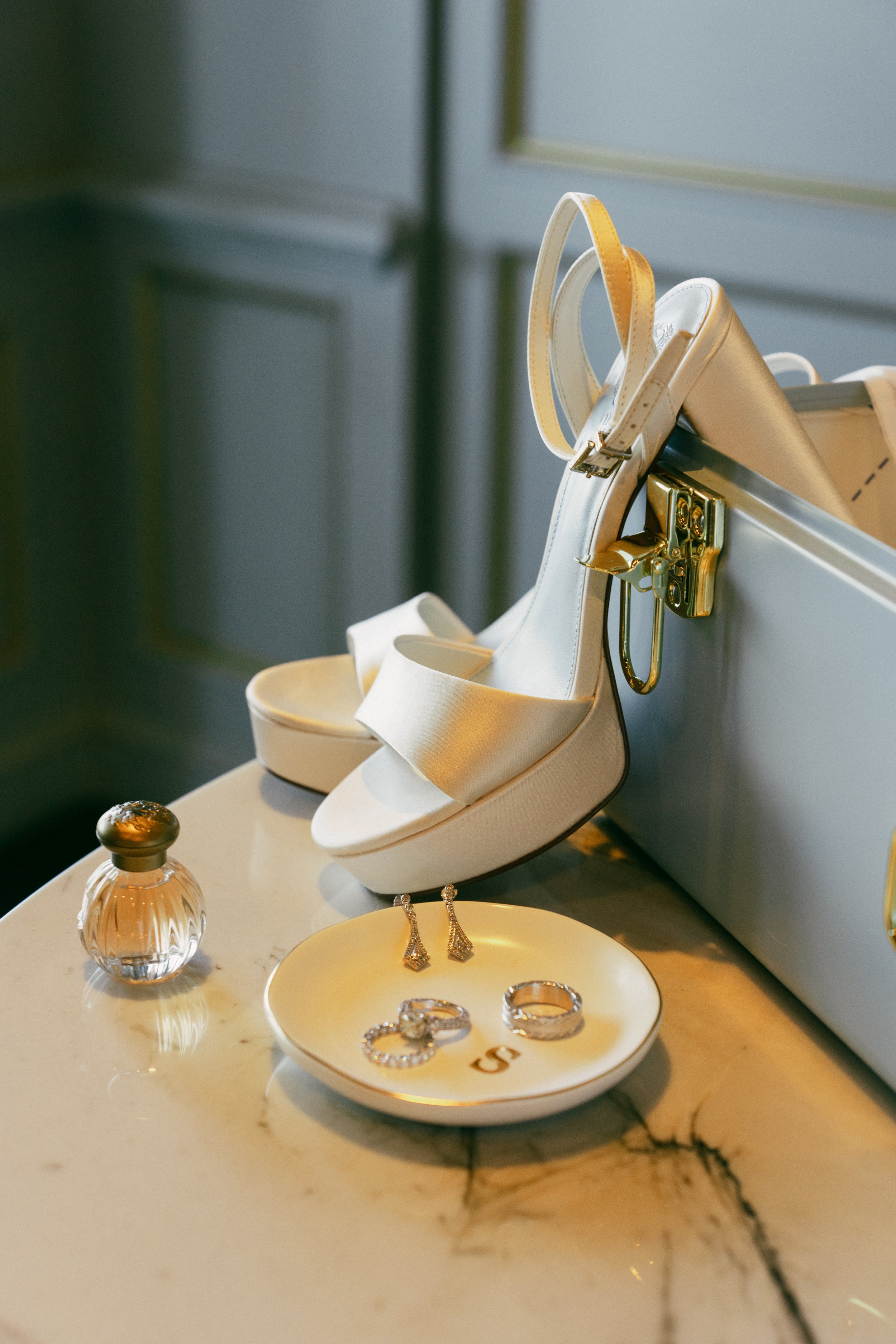 Jewelry display with rings, earrings, and bracelets, along with a white high heel sandal, on a marble surface with a perfume bottle nearby.