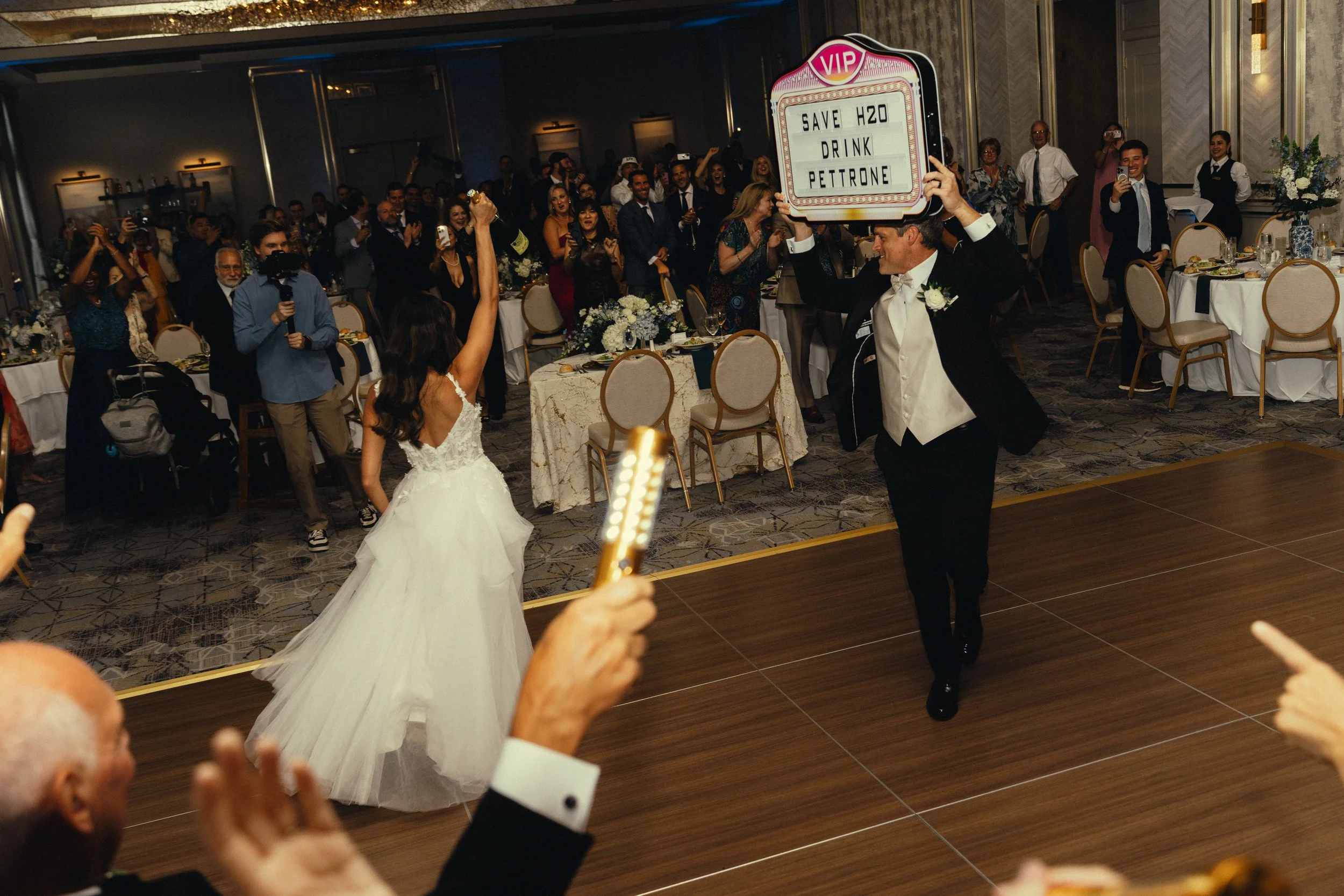 A wedding reception with a bride in a white wedding dress dancing, surrounded by guests seated at decorated tables. A man in a tuxedo holding a sign that says "SAVE H2O, DRINK PETTRONE" is in the center, smiling and dancing. Guests are clapping and t