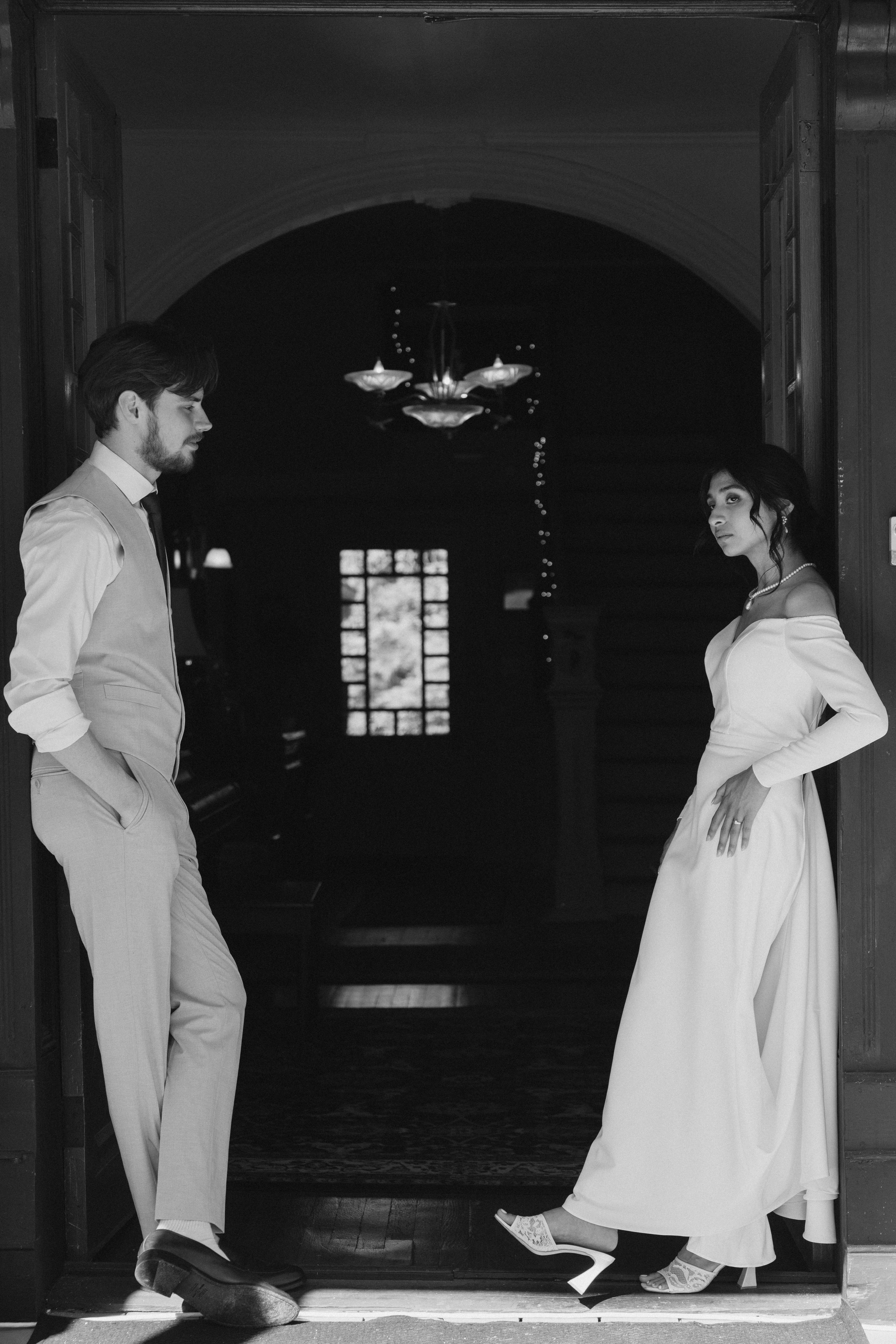 A black and white photo of a man and a woman standing in a doorway inside a building. The man has a beard, styled hair, and is wearing a suit with a vest and tie, with his hands in his pockets, looking down. The woman has dark hair, wearing a straple