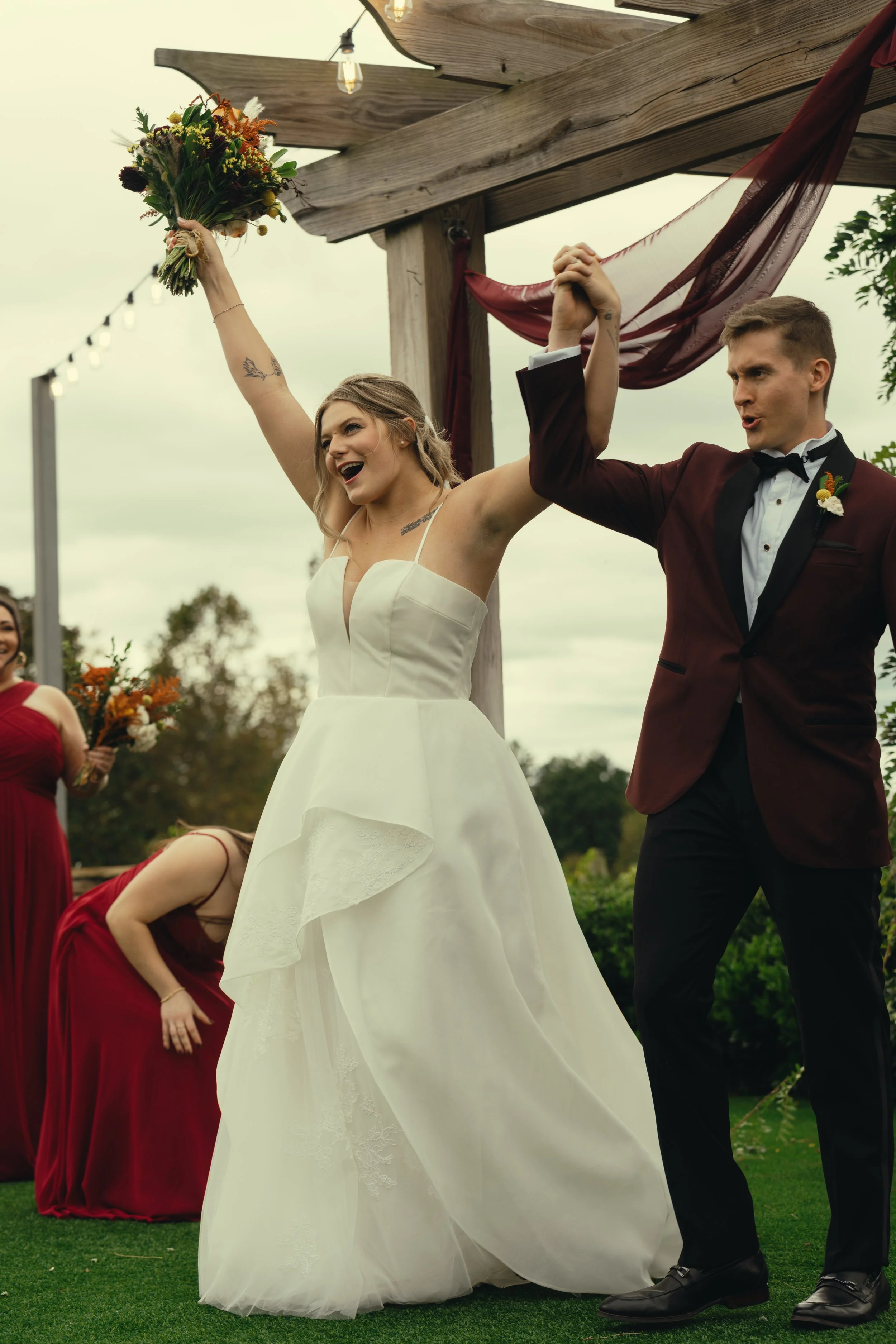 A bride and groom celebrate during their outdoor wedding ceremony, holding hands up in victory. The bride is wearing a white gown and holding a bouquet of flowers, while the groom is wearing a dark tuxedo with a bow tie. There are bridesmaids in red 