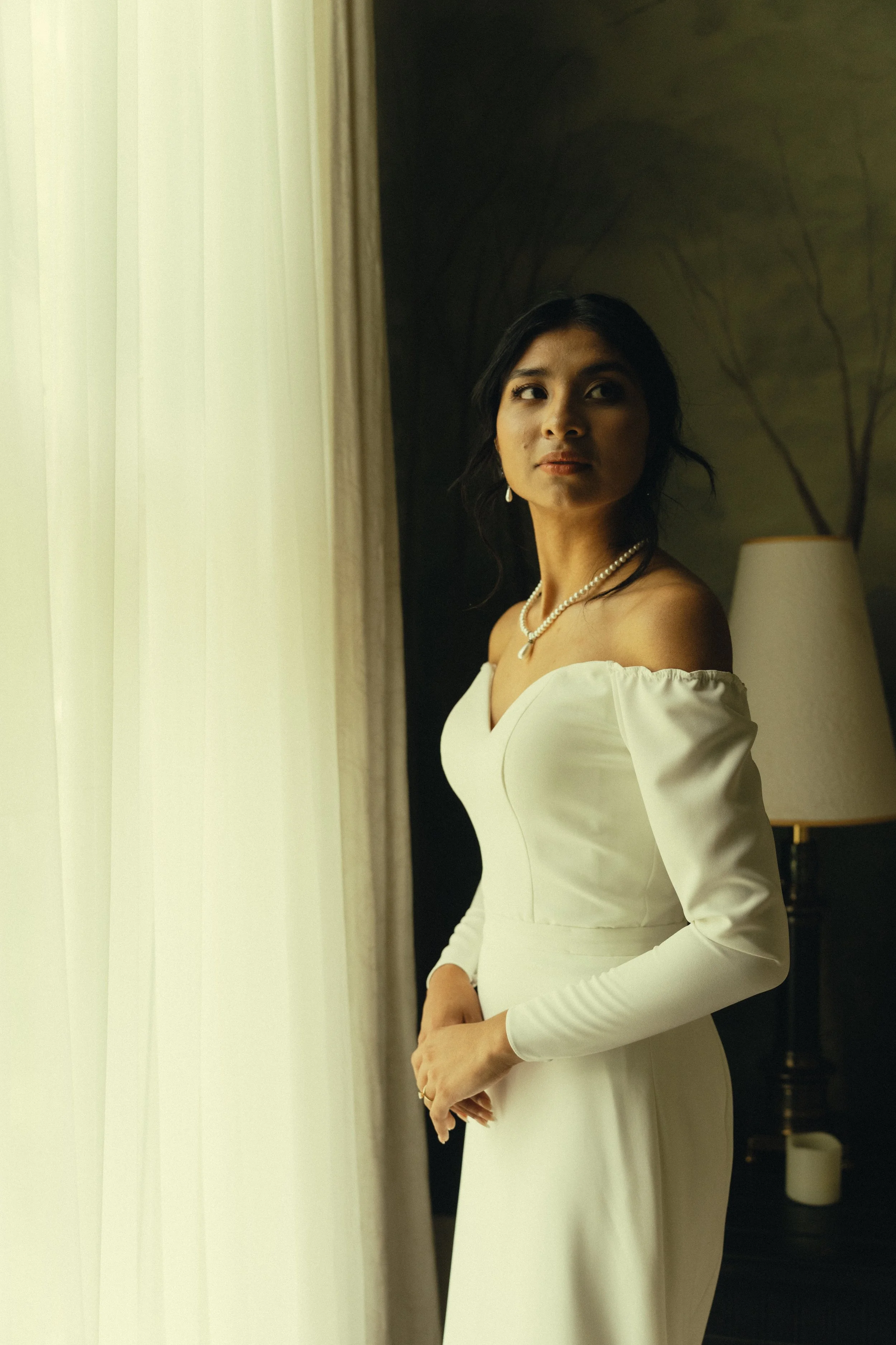 A woman in a white dress standing indoors near a window with curtains, wearing pearl jewelry and looking to the side, with a lamp and a plant silhouette in the background.