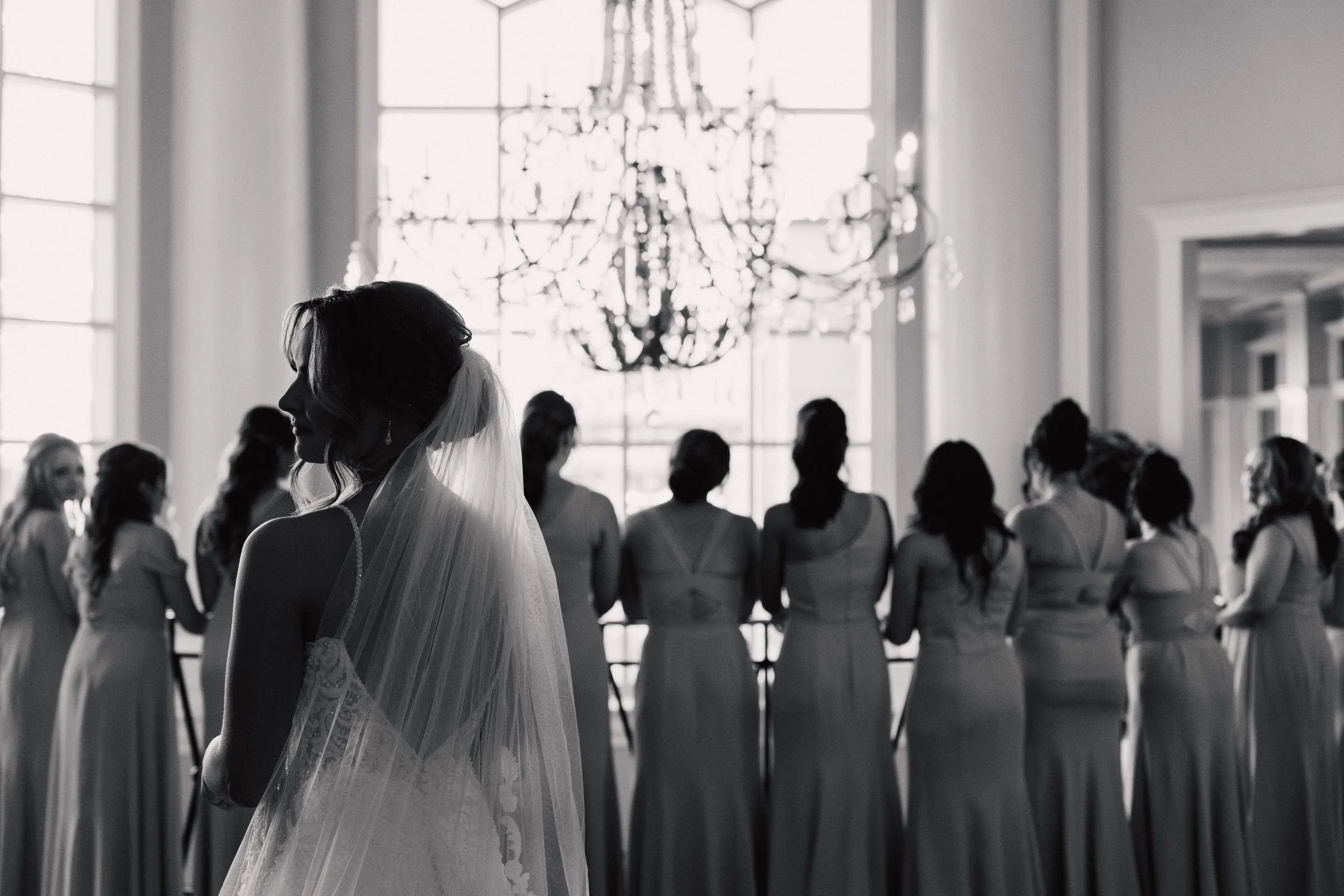 A black and white photo of a bride in a wedding dress standing in front of a group of women, likely bridesmaids, in a large elegant room with a chandelier and tall windows.