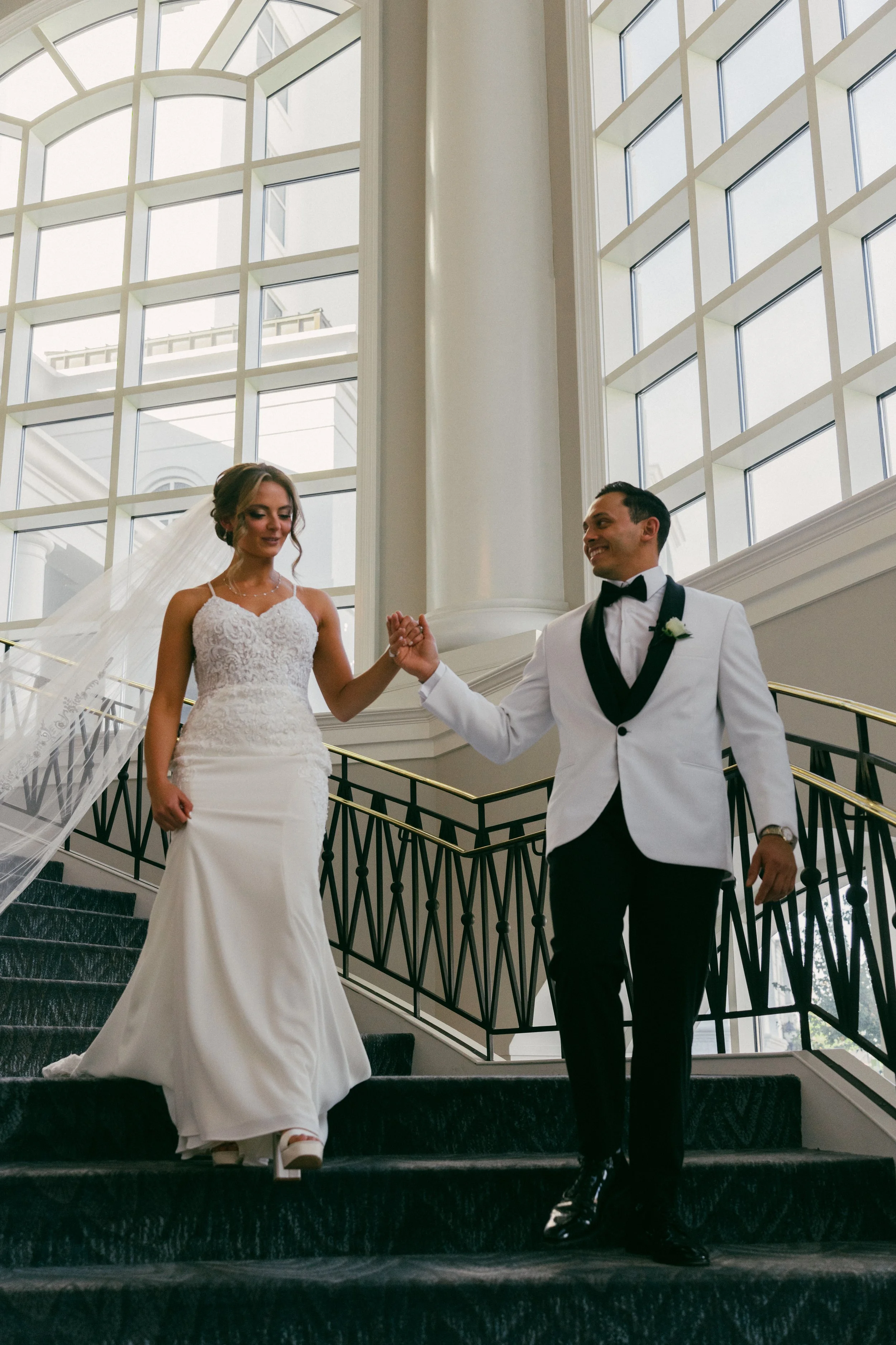 A newlywed couple descending a staircase inside a building with large windows and a grand interior. The bride is wearing a white lace wedding gown and veil, and the groom is dressed in a black and white tuxedo with a bow tie. They are holding hands a