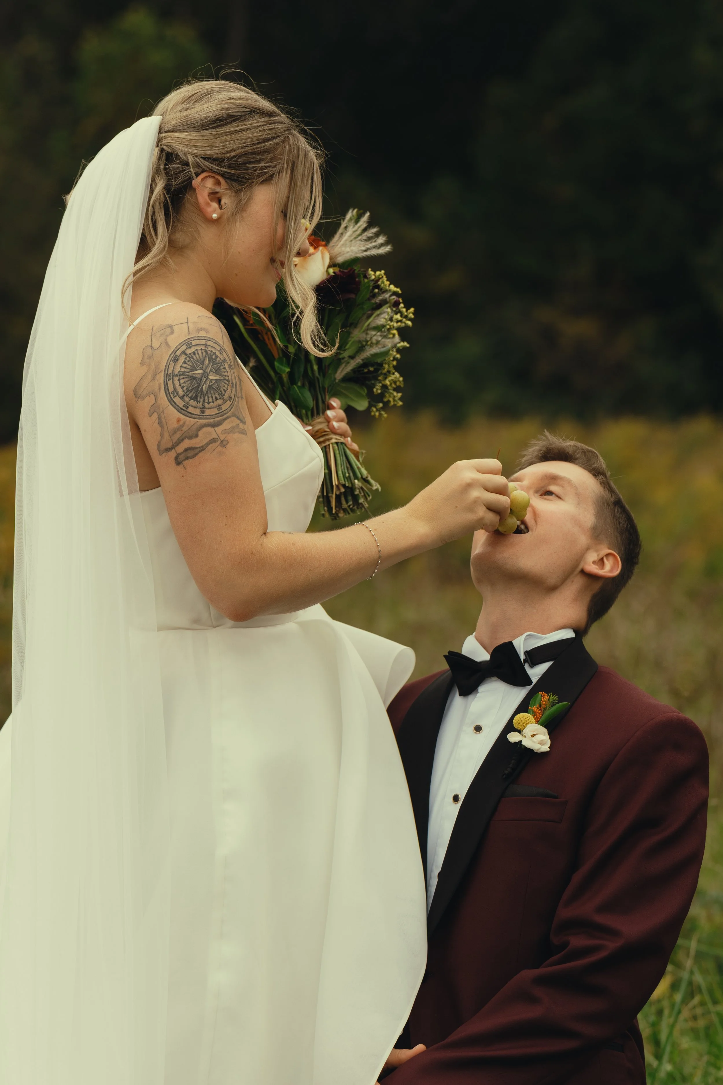 A bride with tattoos on her upper arm holding a bouquet of flowers is feeding a piece of fruit to her groom, who is kneeling and looking up at her. The bride is wearing a white wedding dress and veil, and the groom is dressed in a burgundy tuxedo wit