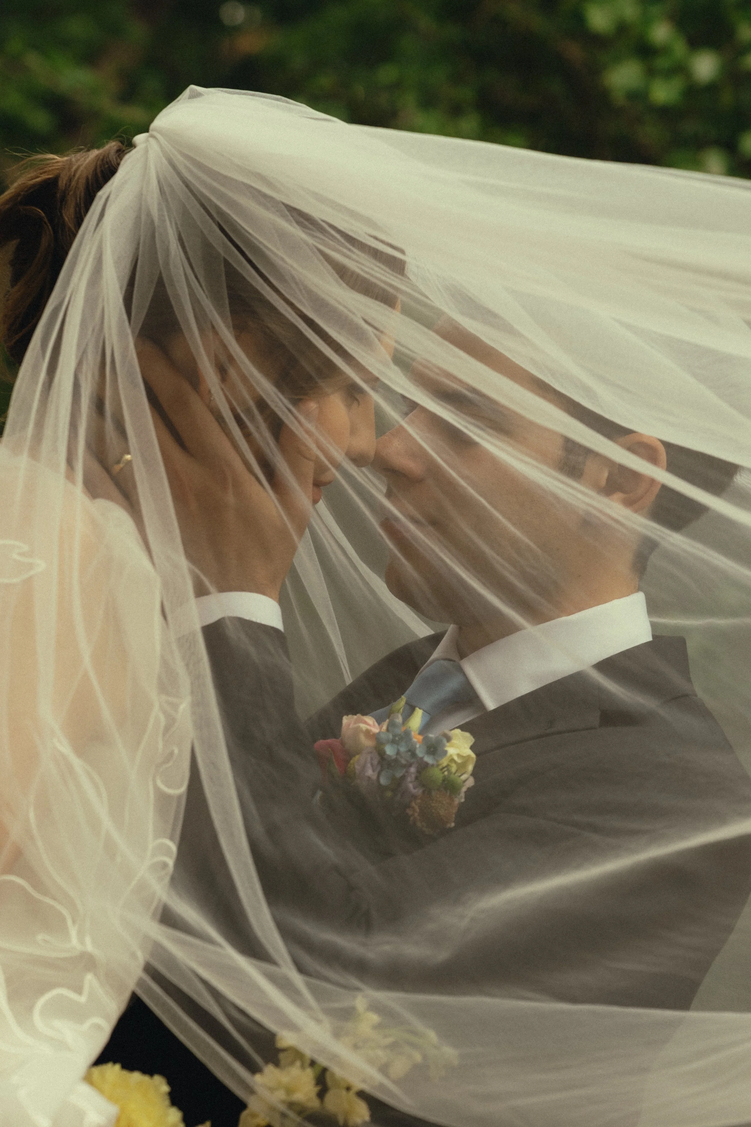 A bride and groom share a kiss outside, with the bride wearing a veil and holding a bouquet of flowers. The groom is in a black suit, and there are greenery and flowers in the background.