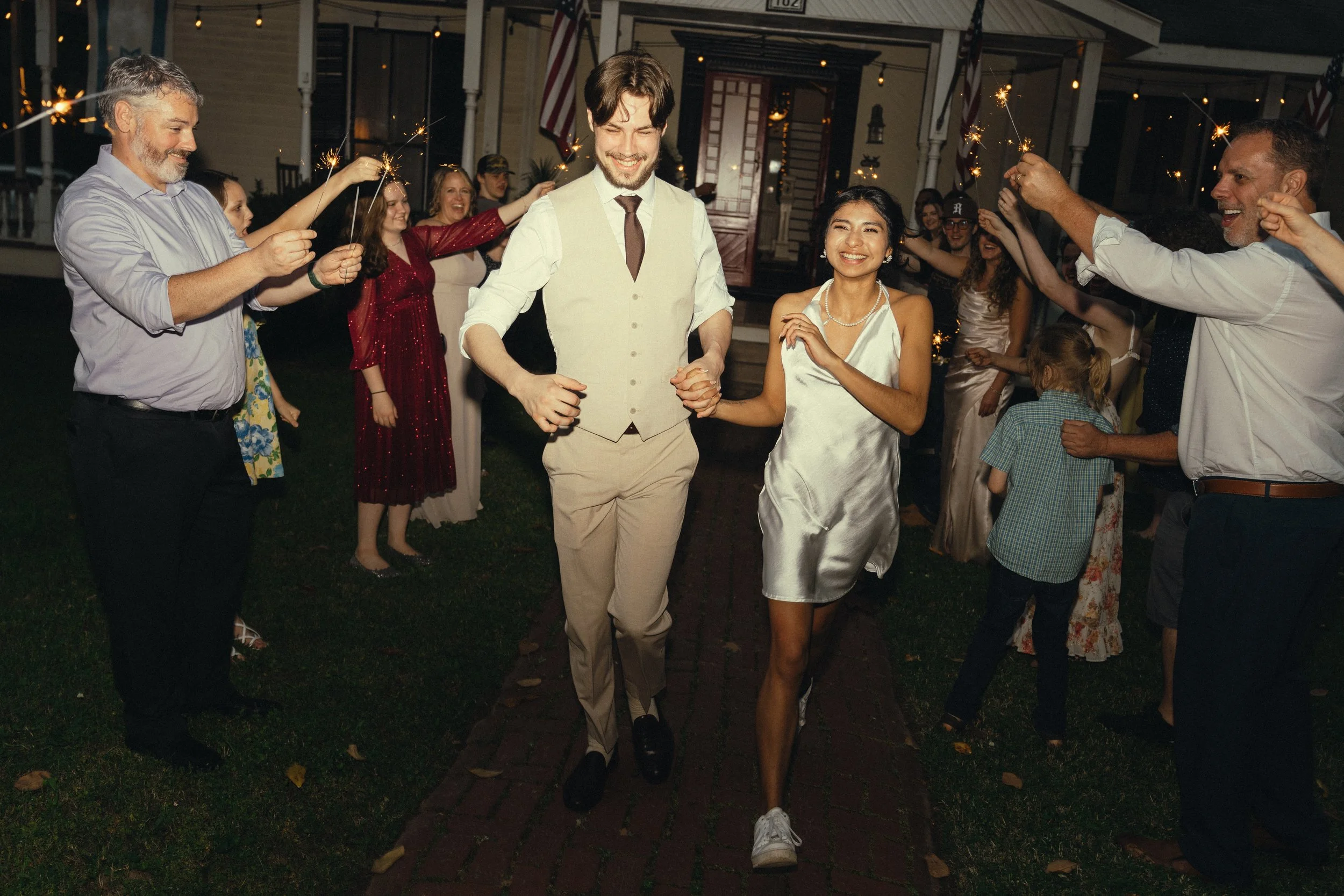 A group of people celebrating outdoors at night, holding sparklers, with a young couple walking hand-in-hand through the crowd, smiling and joyful.