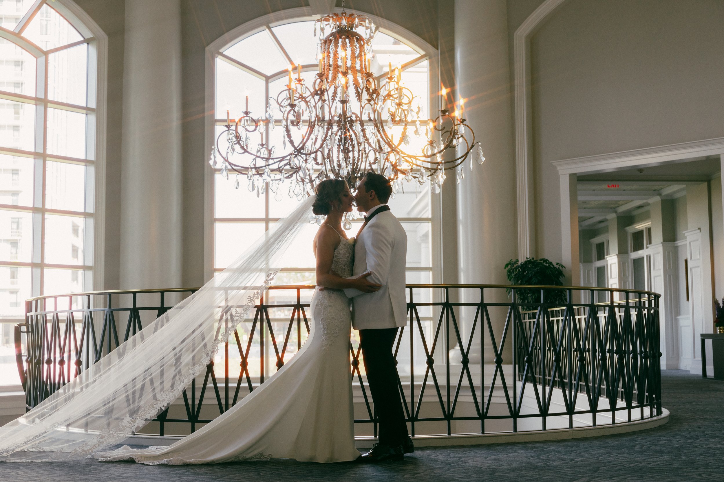 A bride and groom are standing close together in front of a large window with sunlight shining through, during their wedding. The bride is wearing a lace wedding gown and veil, while the groom is dressed in a white tuxedo with a black bow tie. An orn