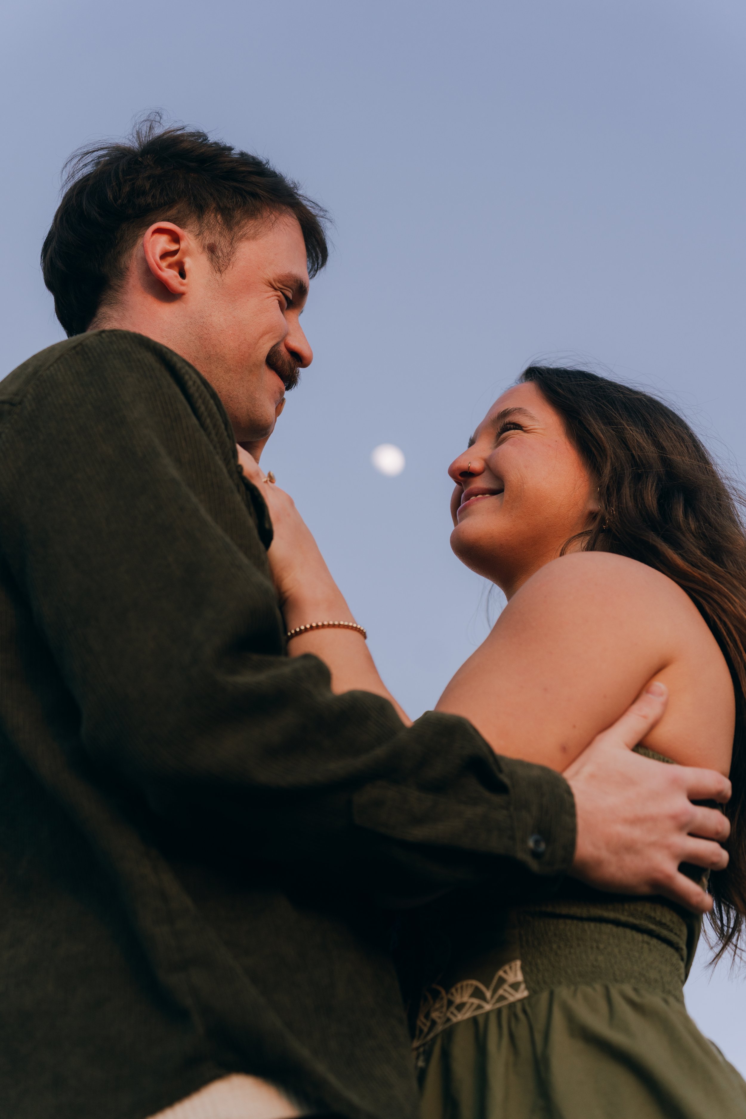 A man and woman smiling and looking at each other, with the moon visible in the background sky.