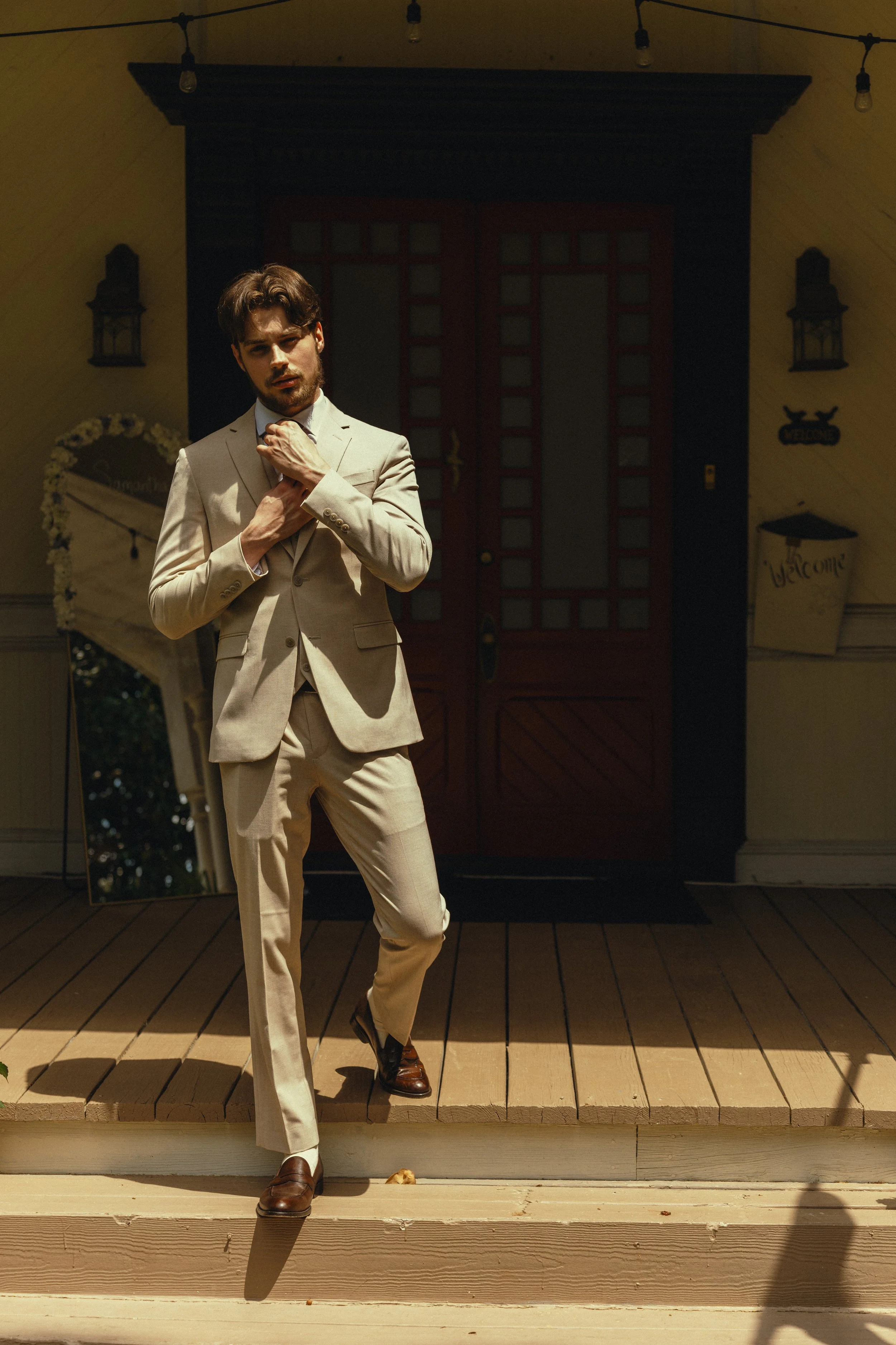 A young man in a beige suit adjusting his tie, standing on a wooden porch in front of a dark red door with grid windows. The porch has wooden steps and a yellow wall with lanterns, a mirror, and decorative welcome signs.