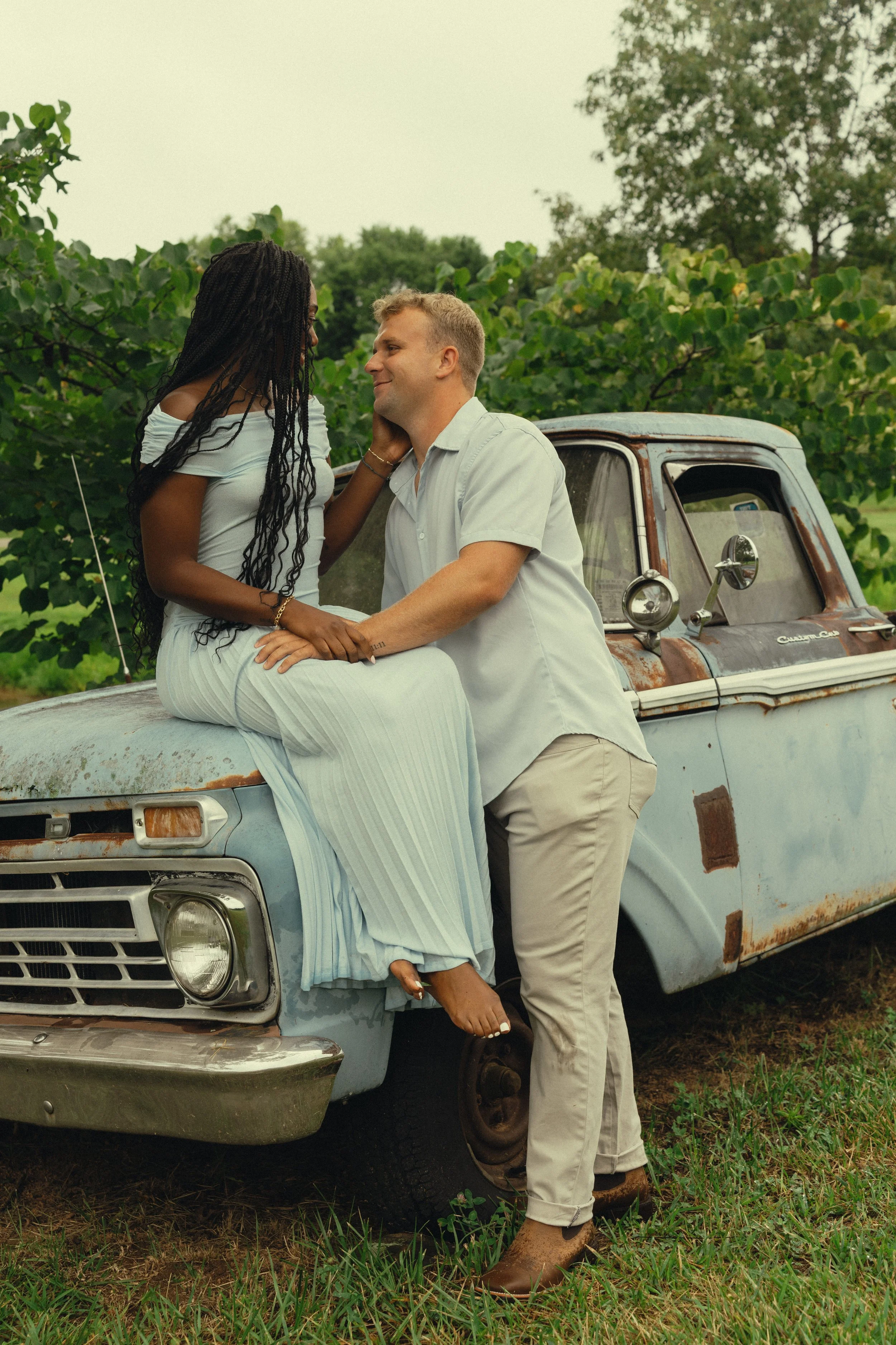 A couple is sitting on the hood of an old rusted blue truck in a field surrounded by greenery and trees, with the woman touching the man's face and both smiling at each other.