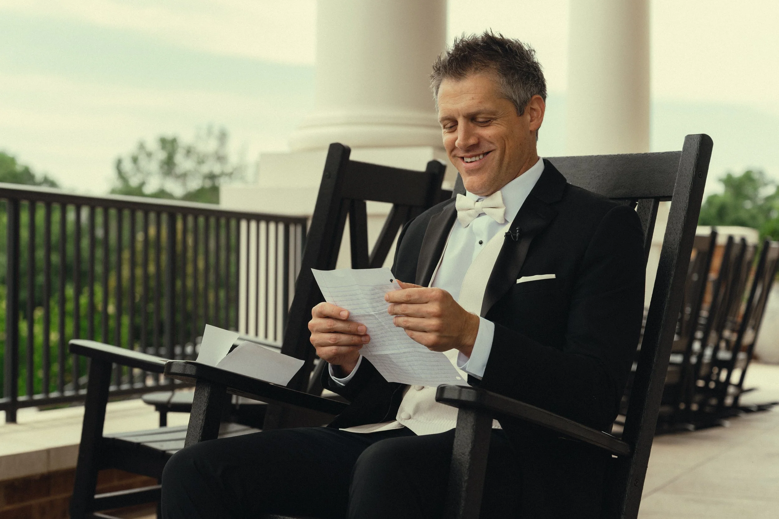 A man in a tuxedo with a white bow tie sitting on a porch chair, smiling while looking at papers in his hands.