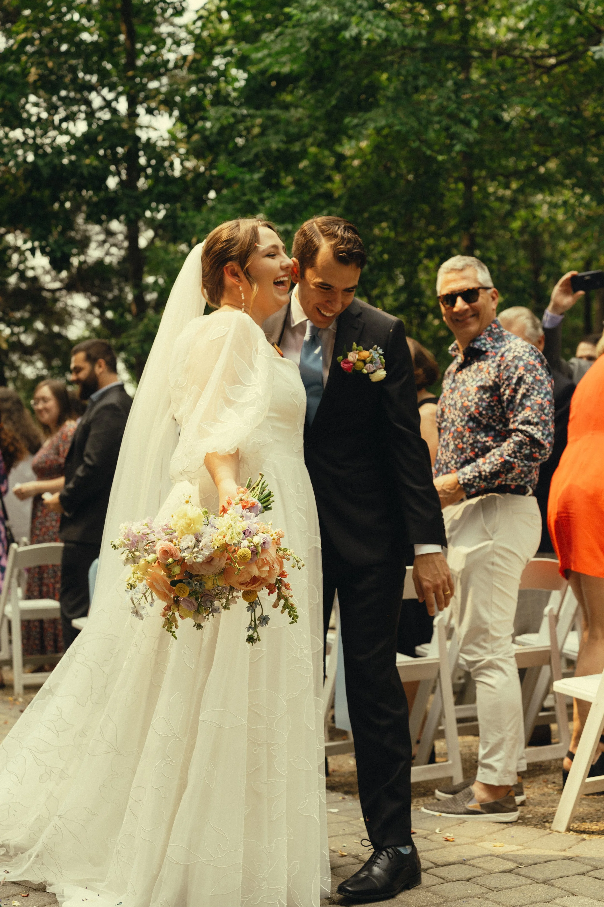 A bride and groom smiling and laughing at their wedding ceremony outdoors with guests in the background. The bride holds a bouquet of flowers.