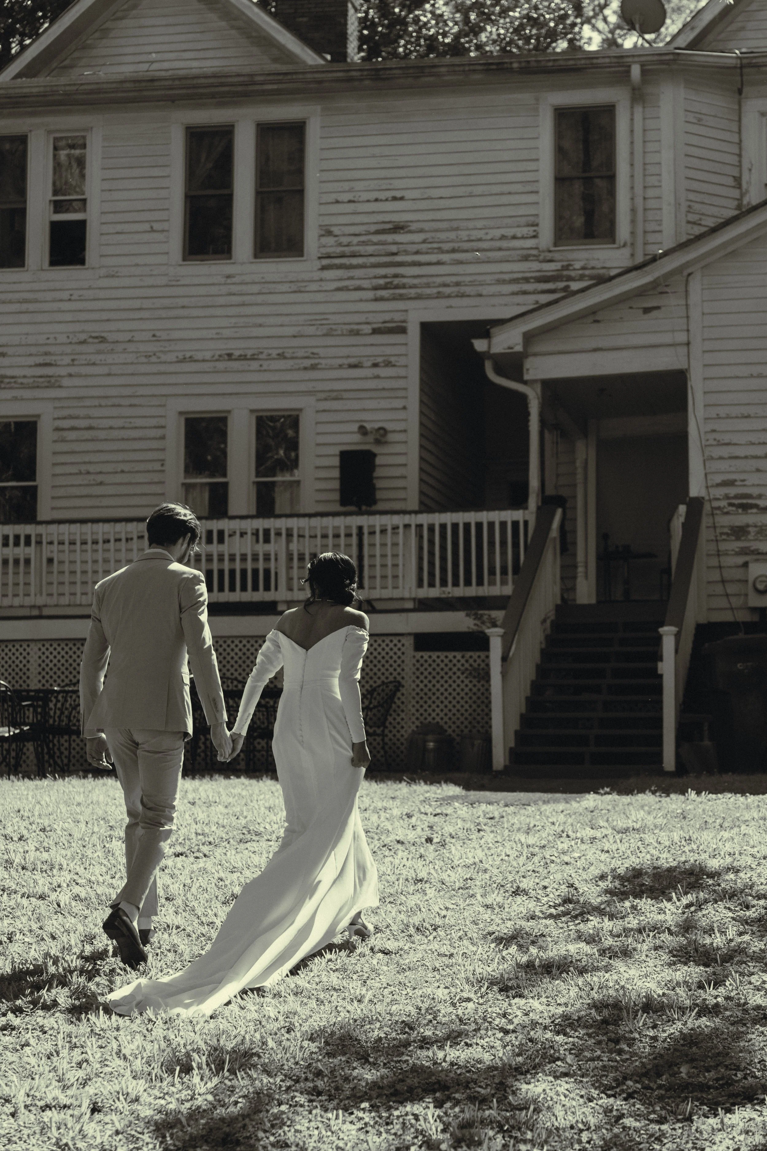 A black and white photograph of a couple walking hand in hand in a backyard, with a weathered two-story house in the background. The man is wearing a light-colored suit, and the woman is in a long, white dress.