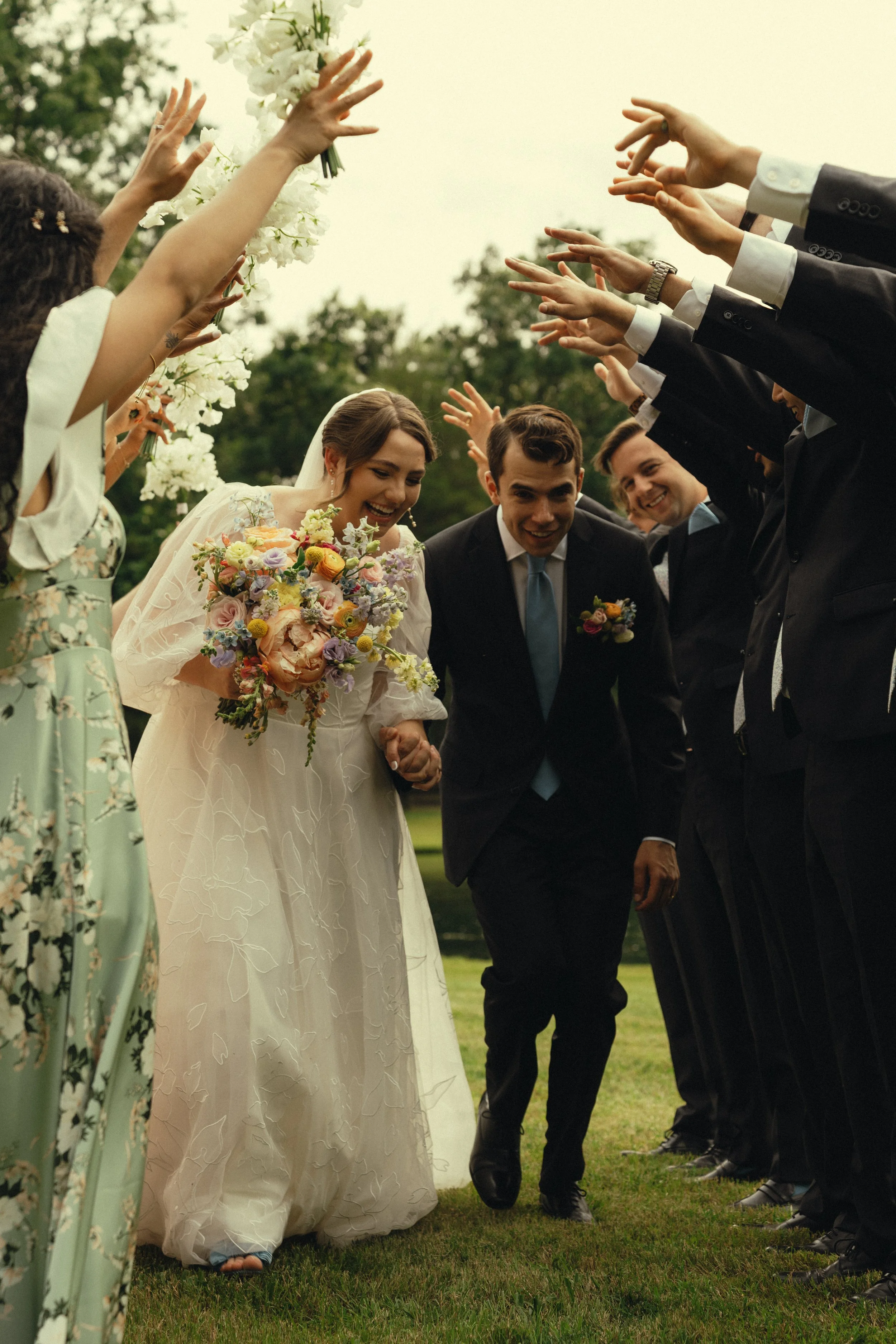 A bride and groom hold hands in a grassy area while surrounded by wedding guests raising their hands with flowers and holding each other in a celebratory moment.