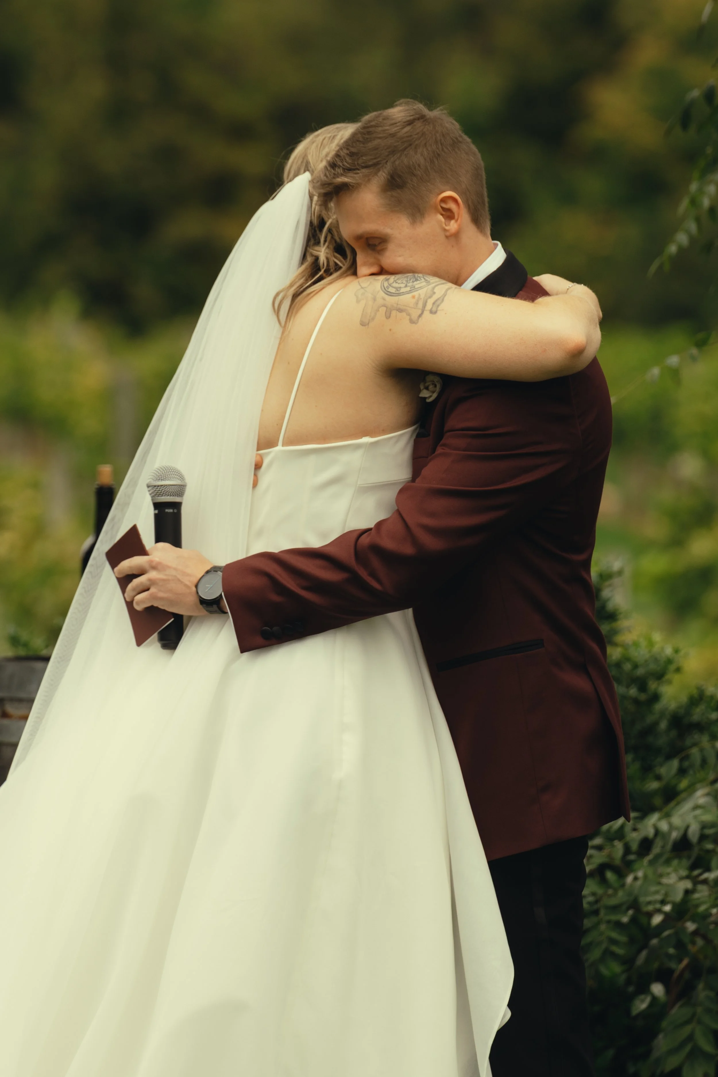 A couple hugging outdoors, woman in a white wedding dress with a long veil, man in a burgundy suit with a black collar, in front of green trees, holding a microphone and a small folder.