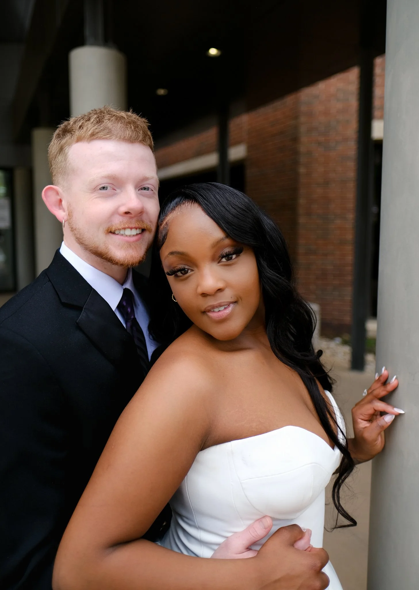 A smiling couple, a man with red hair and a woman with dark hair, posing close together outdoors near a building with brick walls, with the woman touching a gray concrete pillar.