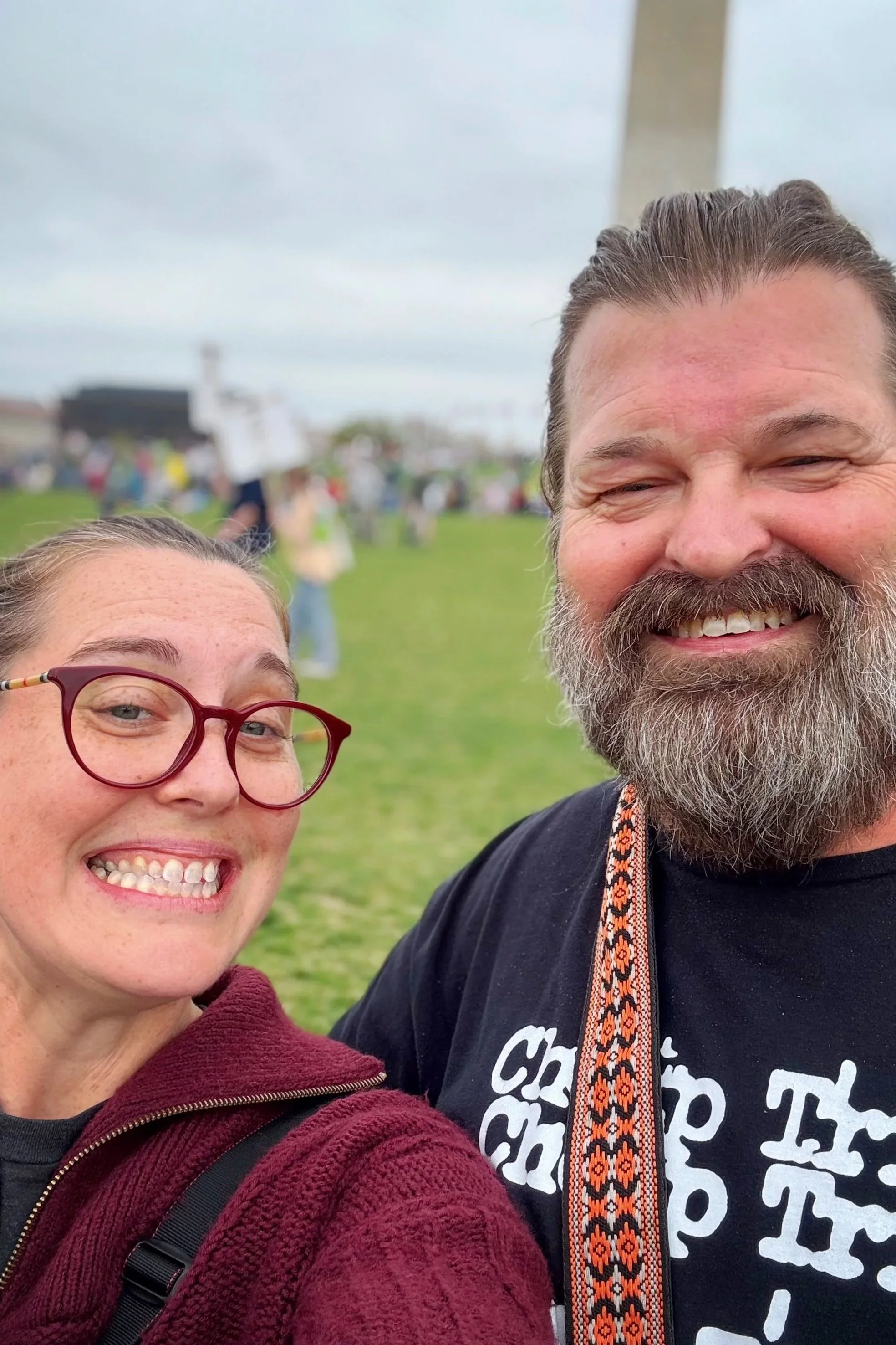 Two people smiling outdoors with a background of a grassy field and cloudy sky. One person is wearing red glasses and a maroon hoodie, and the other is wearing a black shirt with white text and a decorative strap around their neck.