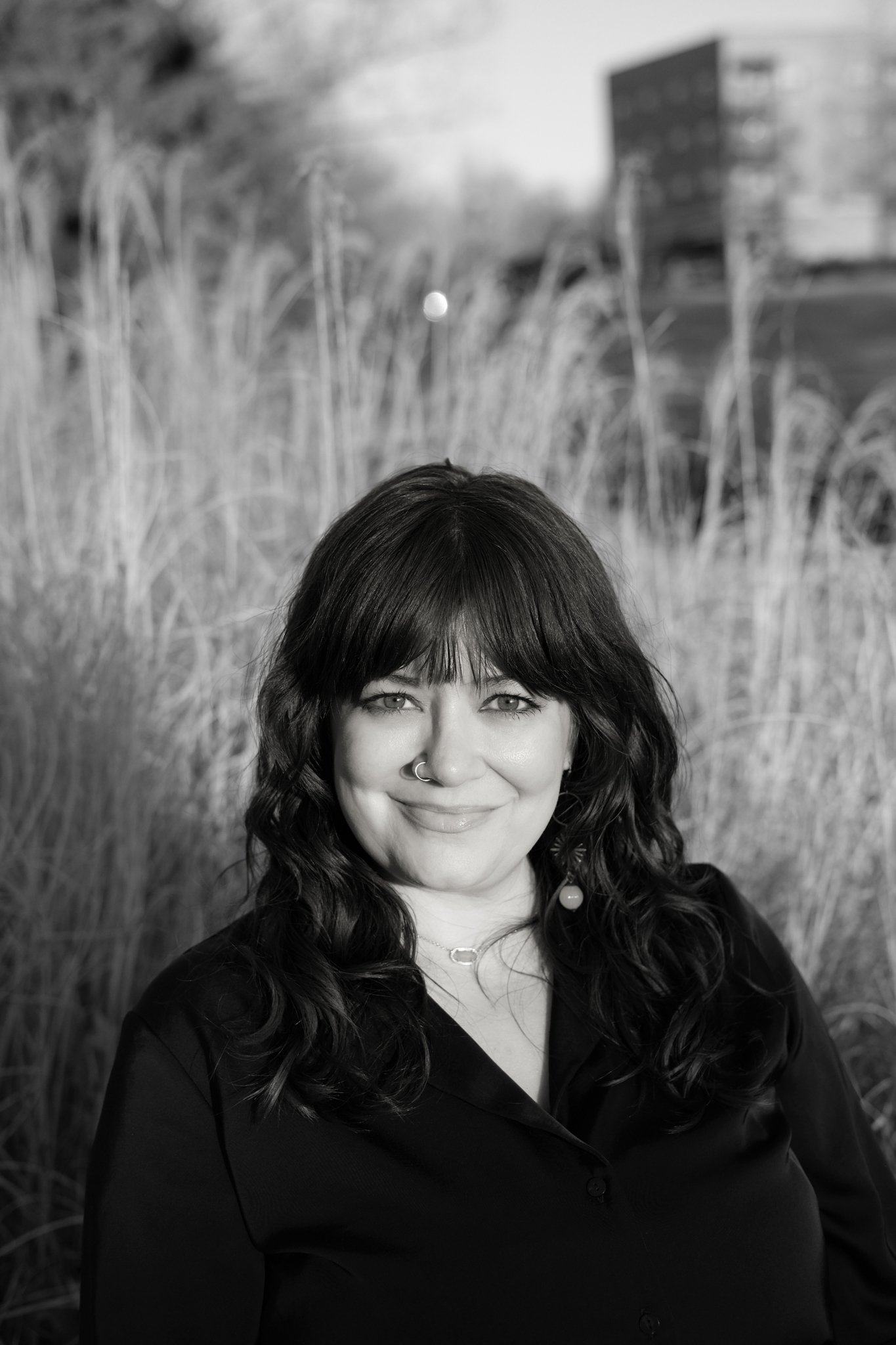Black and white portrait of a smiling woman with long wavy hair, wearing a necklace and a dark top, standing outdoors with tall grasses and buildings in the background.