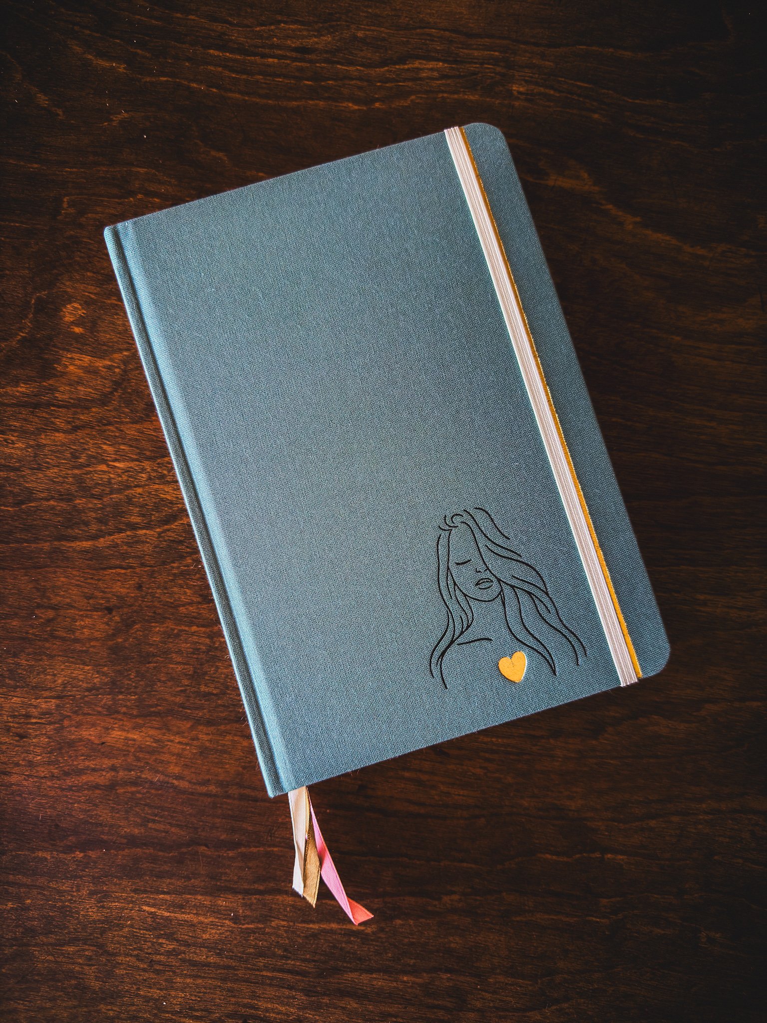 A blue hardcover journal with a line drawing of a woman with closed eyes and flowing hair, a small gold heart, pink and gold ribbons as bookmarks, placed on a dark wooden surface.
