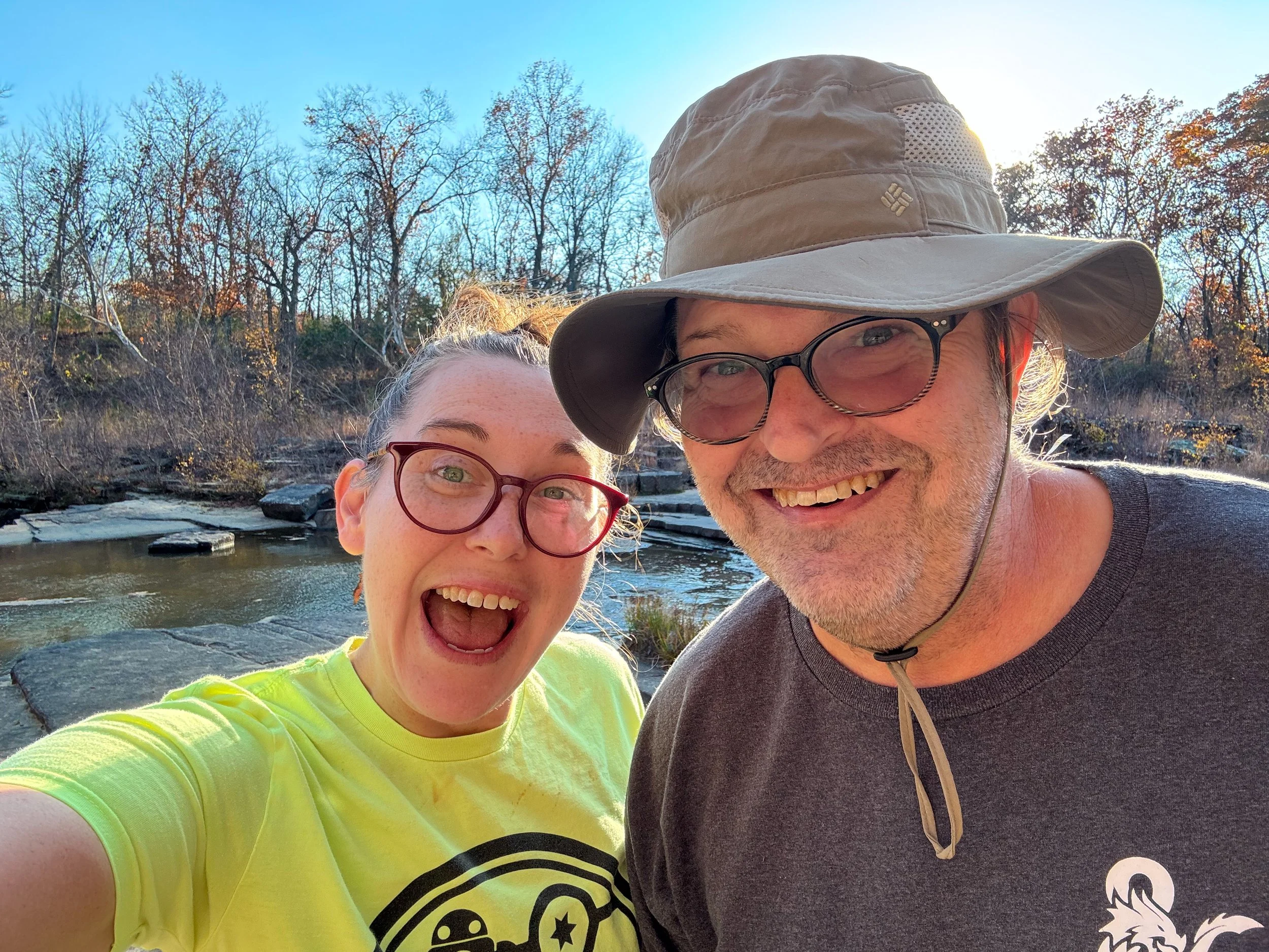 A happy woman with glasses and a yellow shirt and a smiling man with glasses and a wide-brim hat taking a selfie outdoors near a river with rocks and trees with autumn leaves in the background.