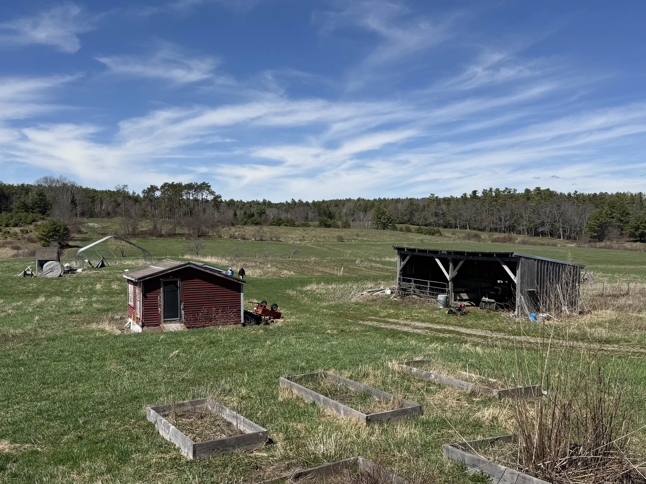A rural landscape with green fields, a few small buildings, and a forested hillside under a mostly cloudy sky.