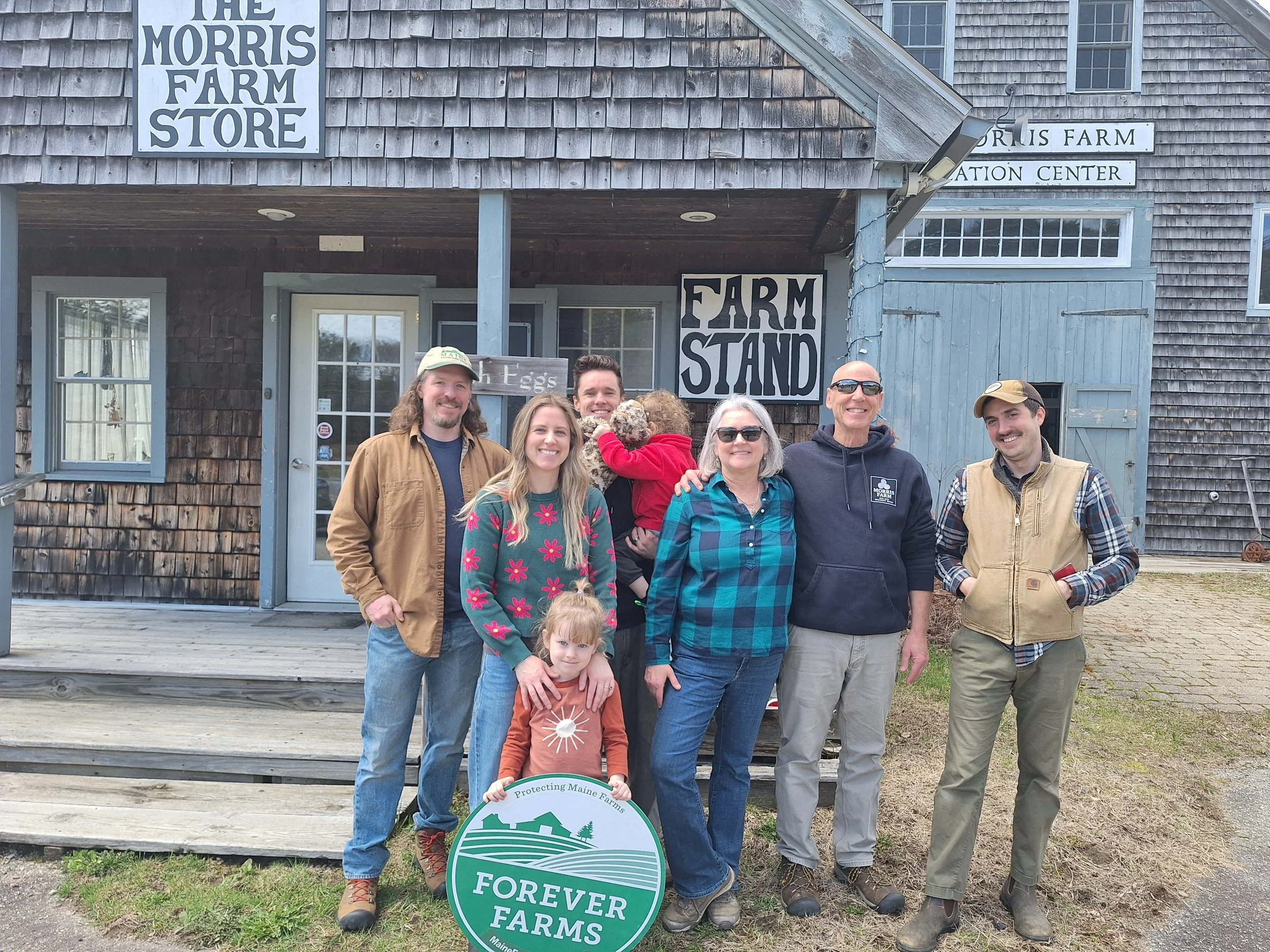 Group of six people smiling in front of a rustic farm building with signs reading "The Morris Farm Store" and "Farm Stand." One person holds a circular sign that says "Forever Farms."