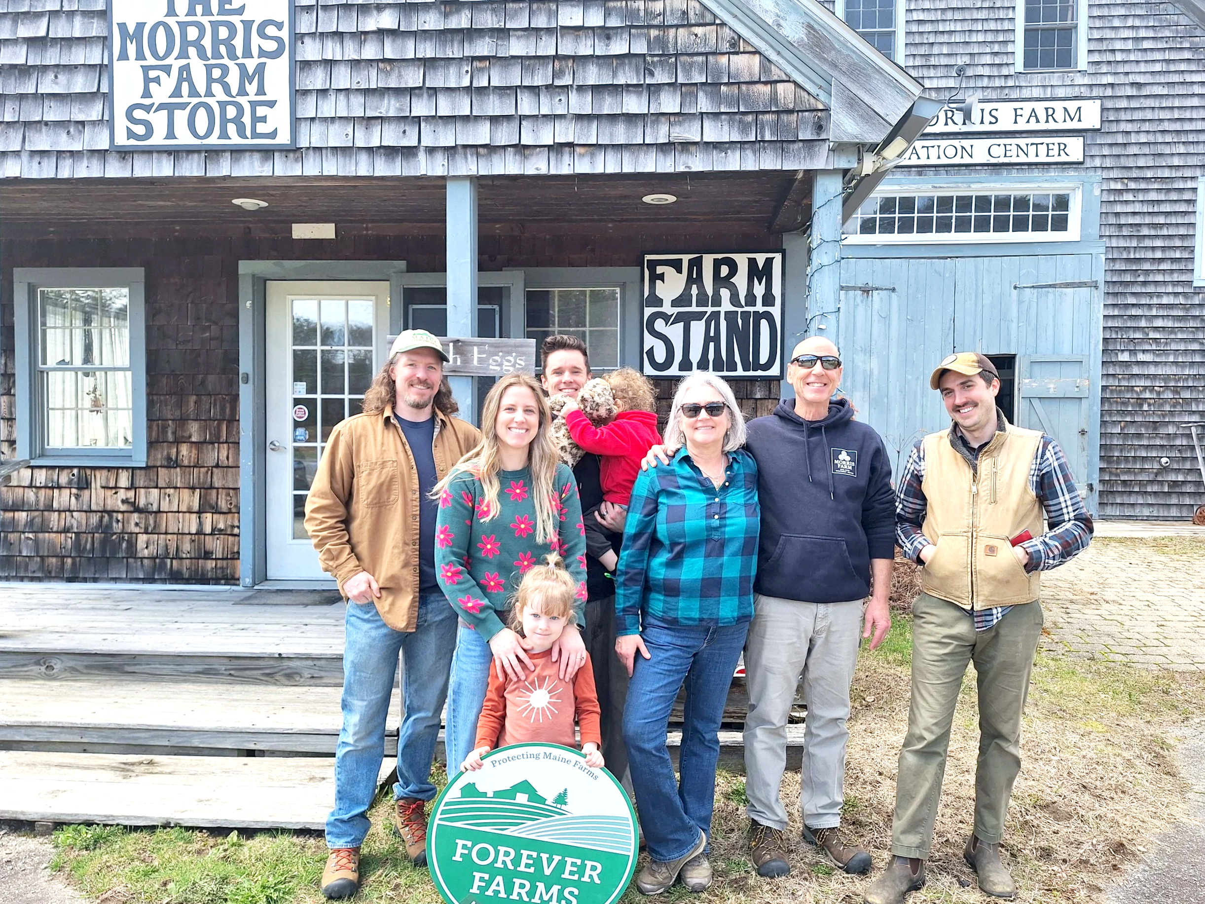 A group of people standing in front of a wooden farm building with signs that say 'The Morris Farm Store,' 'Farm Stand,' and 'Farming Center.' Some are smiling, wearing casual outdoor clothing, and holding farm-related items. One child is in front holding a sign for 'Forever Farms.'