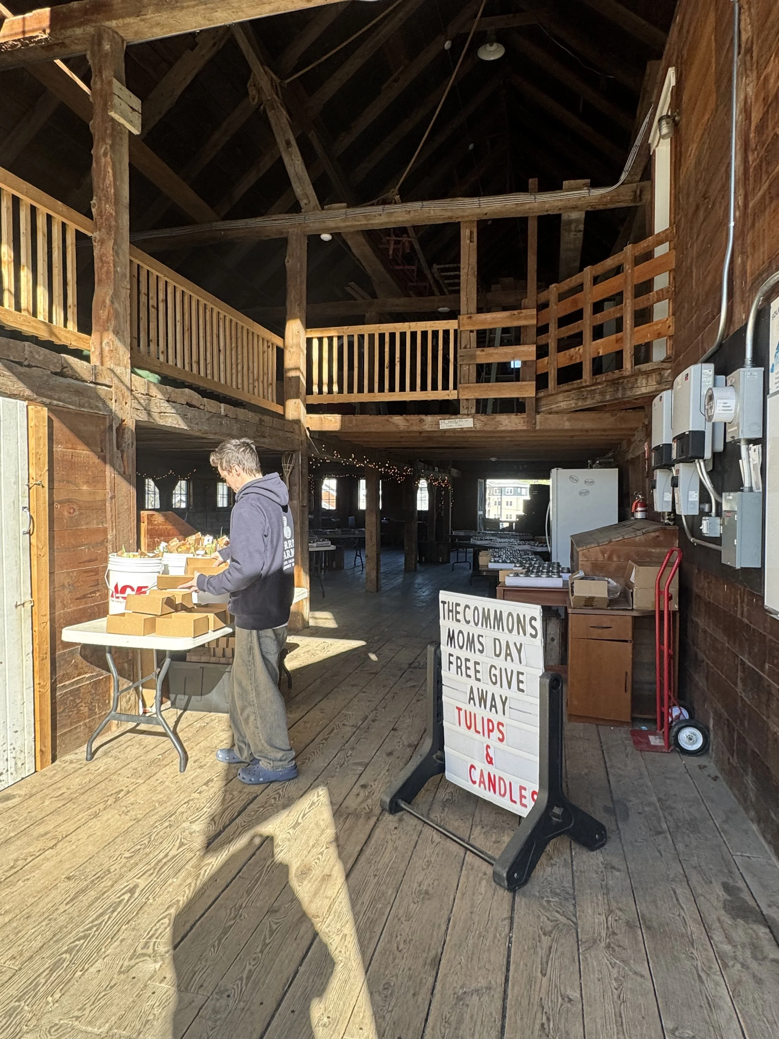A person in a hoodie and Crocs checks out items at a table inside a rustic wooden building with a second-floor balcony. A sign advertises free gifts of flowers and candles for moms on Mother's Day, and a white sign with black letters is visible.