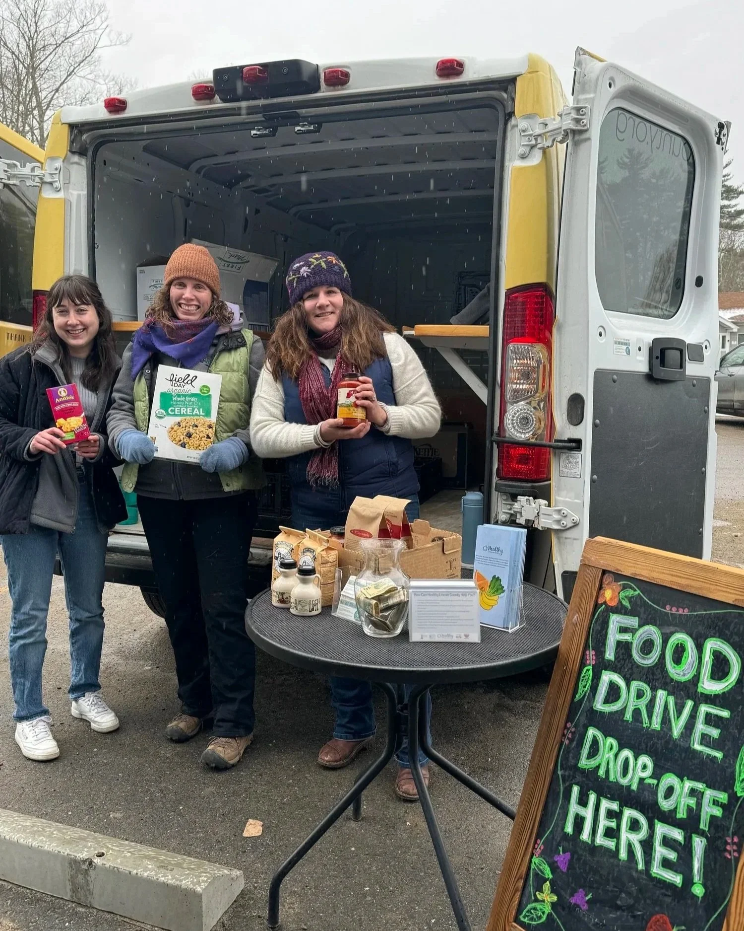 Three women standing in front of a food truck at a food drive event, holding food items, with a table and a chalkboard sign that reads 'Food Drive Drop-Off Here' nearby.