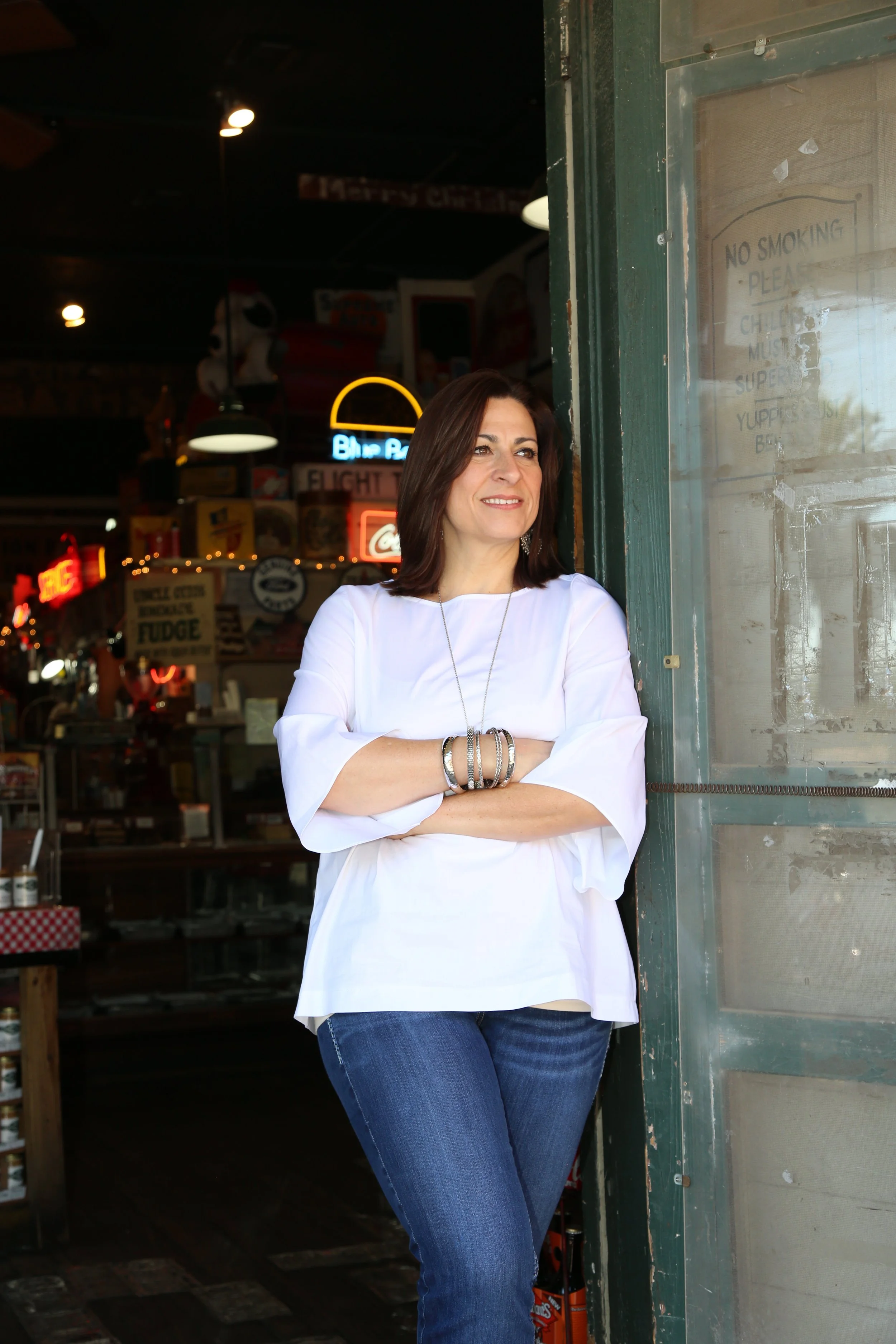 A woman with shoulder-length dark brown hair, wearing a white long-sleeve top and blue jeans, stands with her arms crossed outside a rustic building with a glass door, smiling and looking to the side. The background shows neon signs and a dimly lit interior with various signs and decorations.