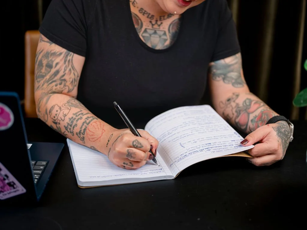 En person med tatoveringer skriver i en åpen notatbok med penn, sitter ved et svart bord.