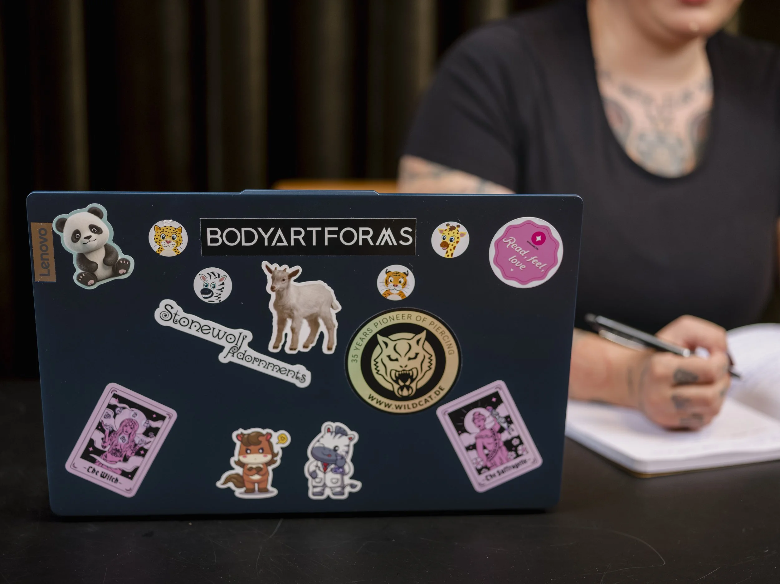 Nærbilde av en laptop dekket av ulike fargerike klistremerker med dyr, tegneserier og tekst, delvis skjult av en person som bruker den.