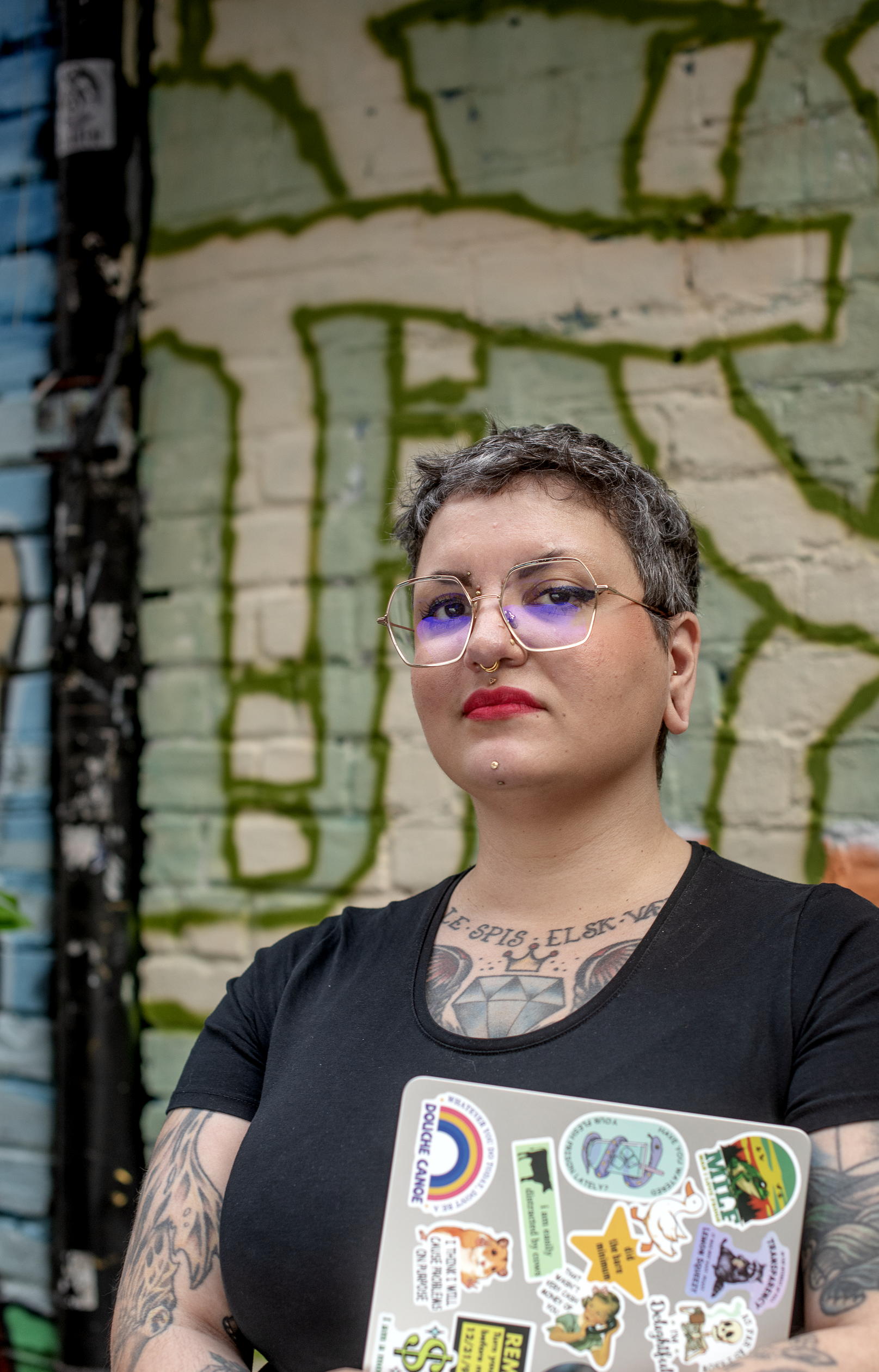 Et portrett av en kvinne med kort grått hår, briller med blå linser, tatoveringer på armer og bryst, som holder en laptop dekket av ulike fargerike klistremerker, foran en graffiti-vegg.