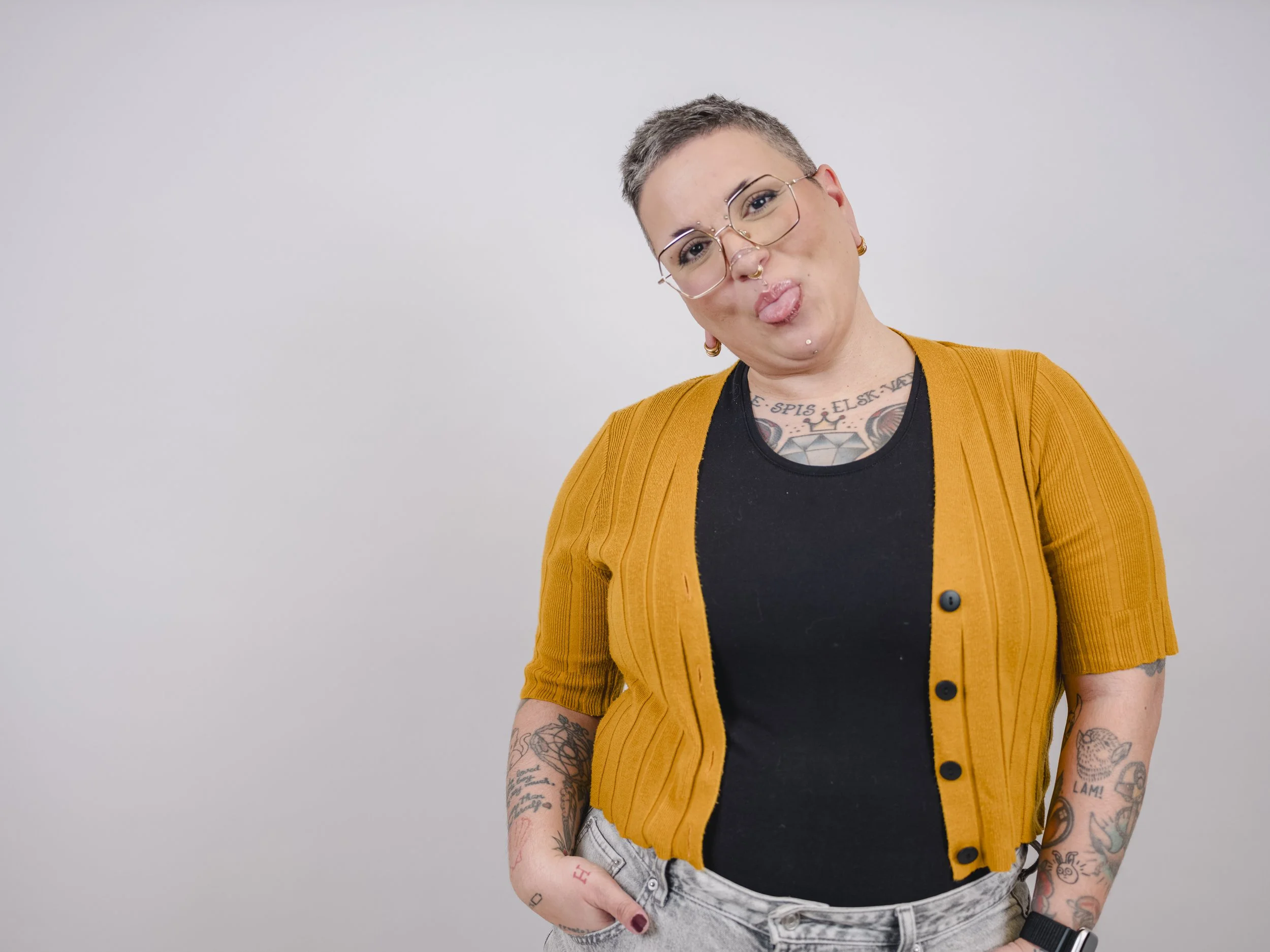 Person med kort grått hår, briller og flere piercinger gjør en tøysete ansiktsuttrykk med tungen ut. De har tatoveringer på armene, definert i en svart t-skjorte og en gul cardigan, og står foran en enkel grå bakgrunn.