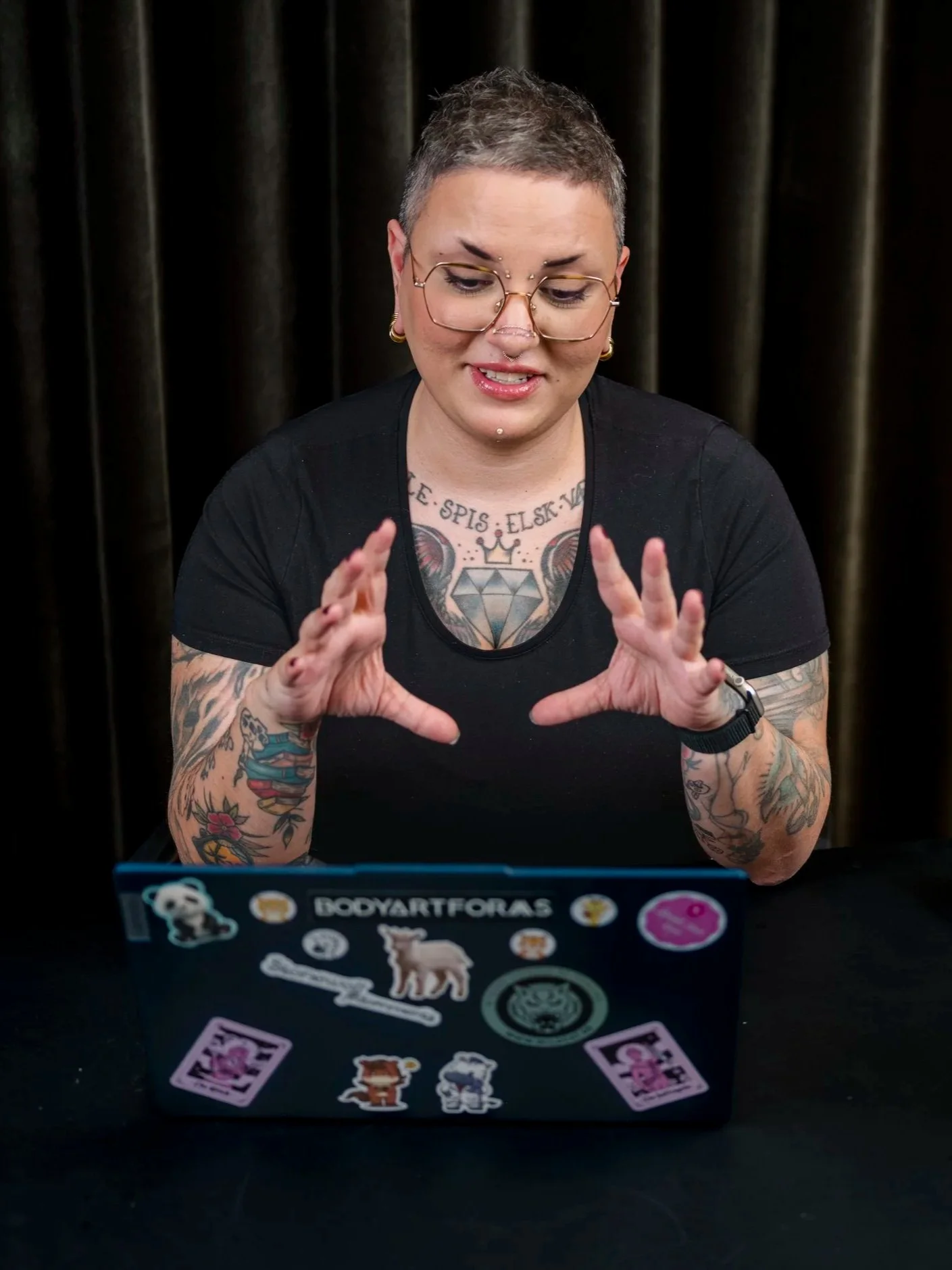 Person med kort, grått hår og flere tatoveringer på armene, som sitter foran en åpen bærbar datamaskin dekorert med ulike klistremerker. Person med briller, smykker og svart t-skjorte gestikulerer mot skjermen.