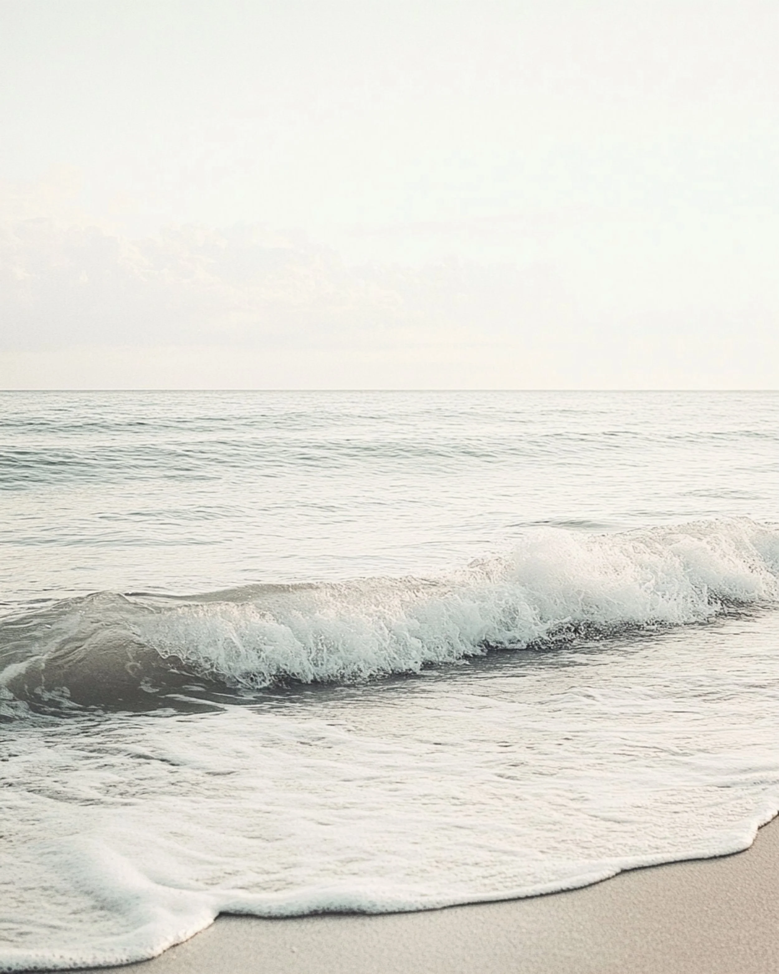 Waves gently rolling onto a beach with light-colored sand, calm water, and a pale sky.