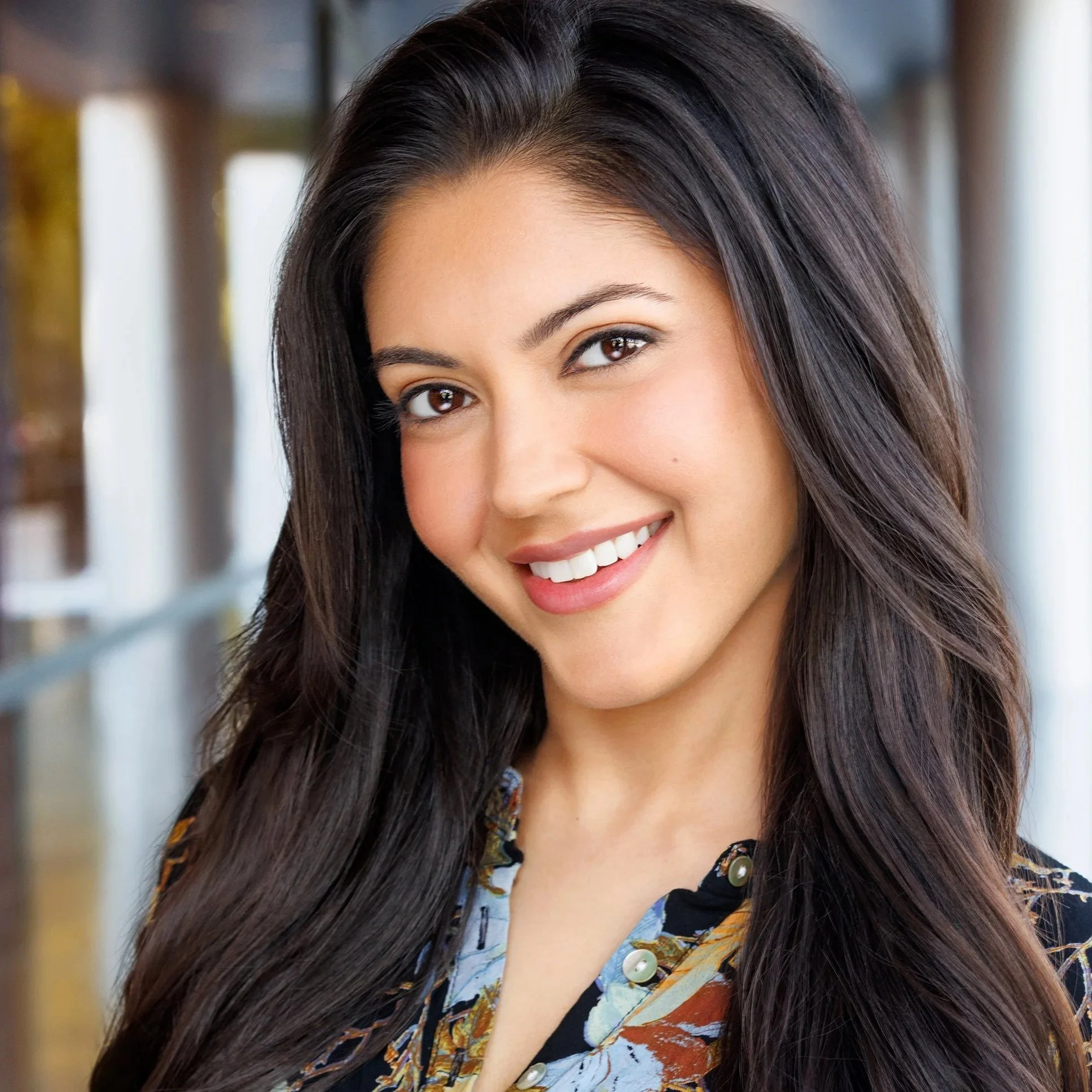 Close-up of a smiling woman with long, dark hair. She has brown eyes and is wearing a colorful, patterned blouse with buttons.