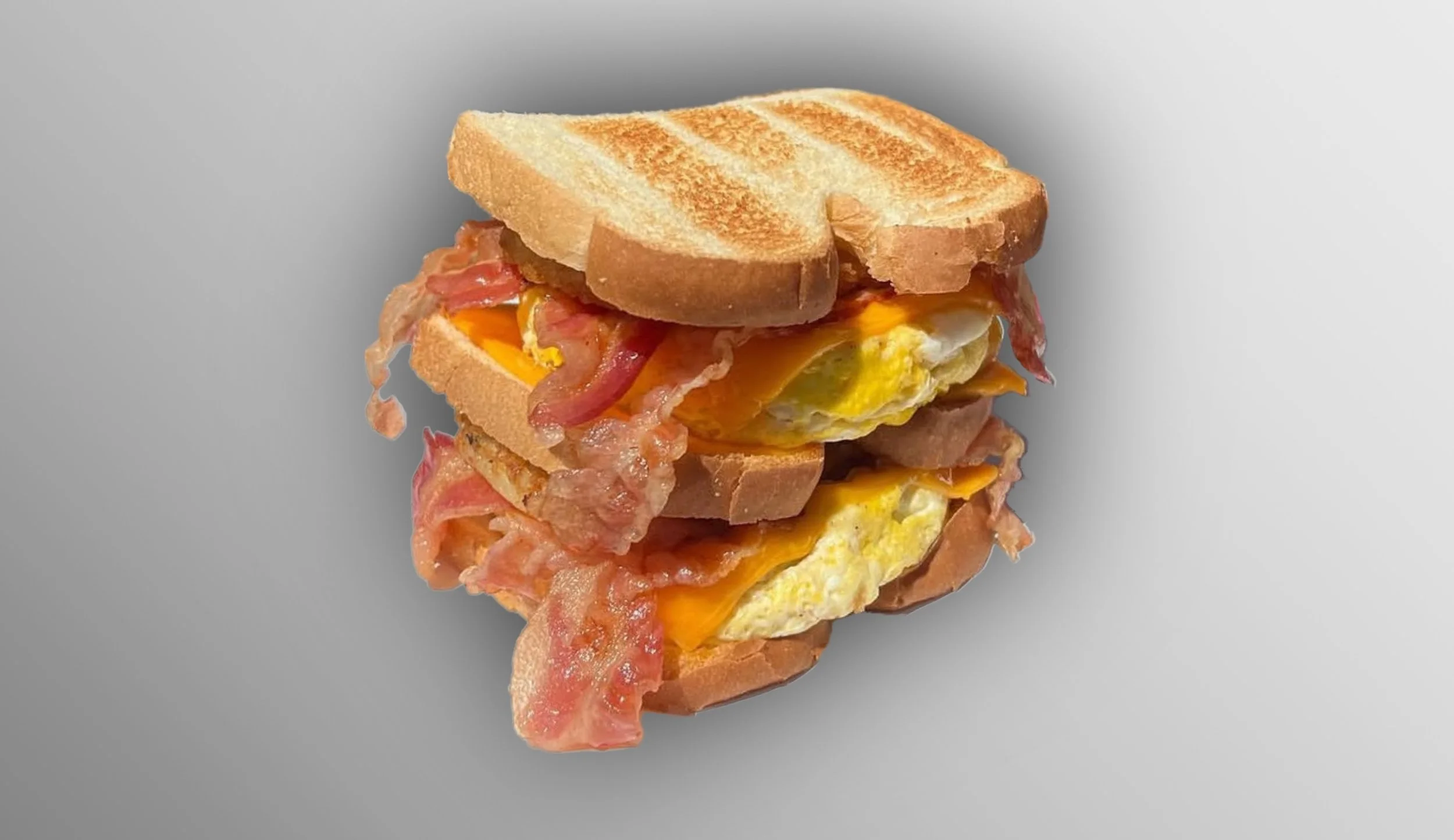 Stacked breakfast sandwich with toasted bread, bacon, melted cheese, and scrambled eggs on a gray background.