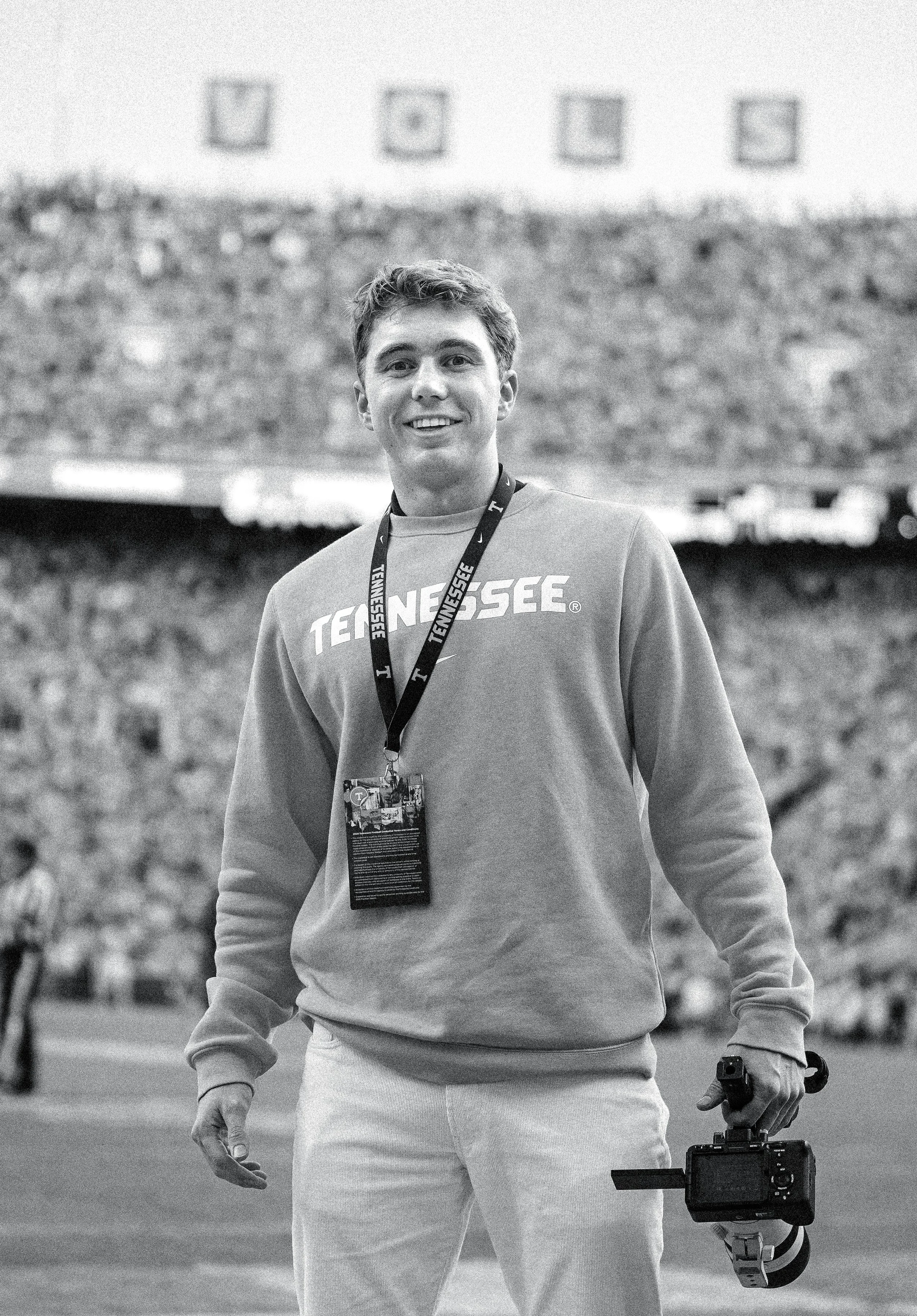 Black and white photo of a young man smiling, wearing a Tennessee sweatshirt, with a lanyard and badge around his neck, holding a camera stabilized by a gimbal, standing outdoors in front of a wall with abstract artwork.