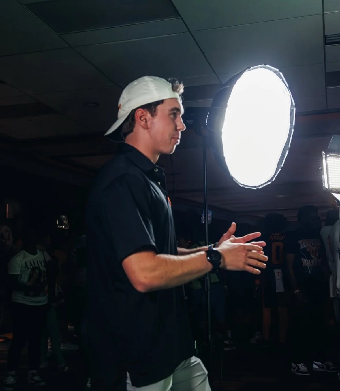 Young man wearing a white cap backwards and a black polo shirt, standing in front of a bright light, possibly in a recording studio or event space, gesturing with his hands.