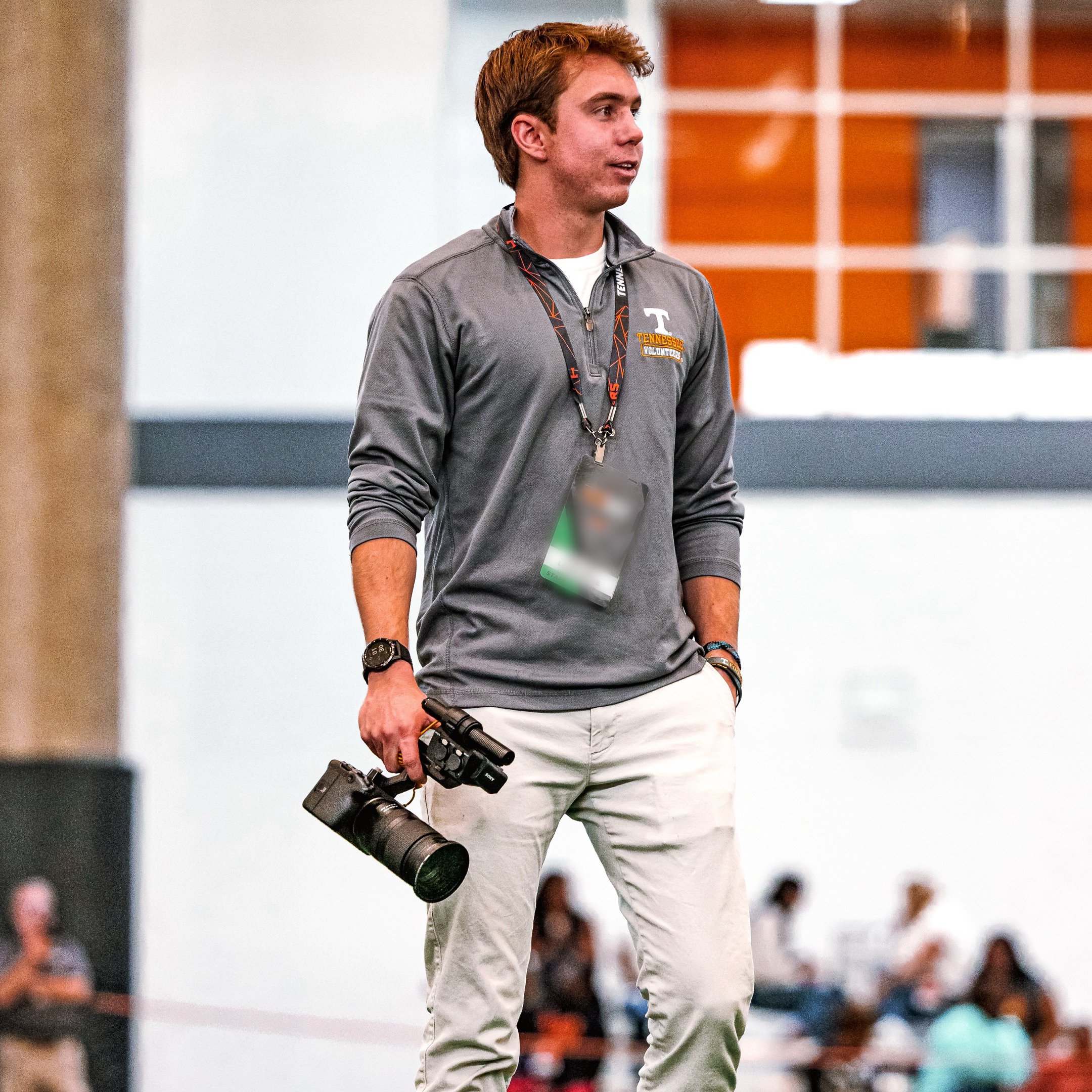 A young man with light skin and light brown hair holding a large camera lens, wearing a gray jacket with a 'Tennessee Volunteers' logo, standing in a gymnasium or sports arena with blurred spectators in the background.