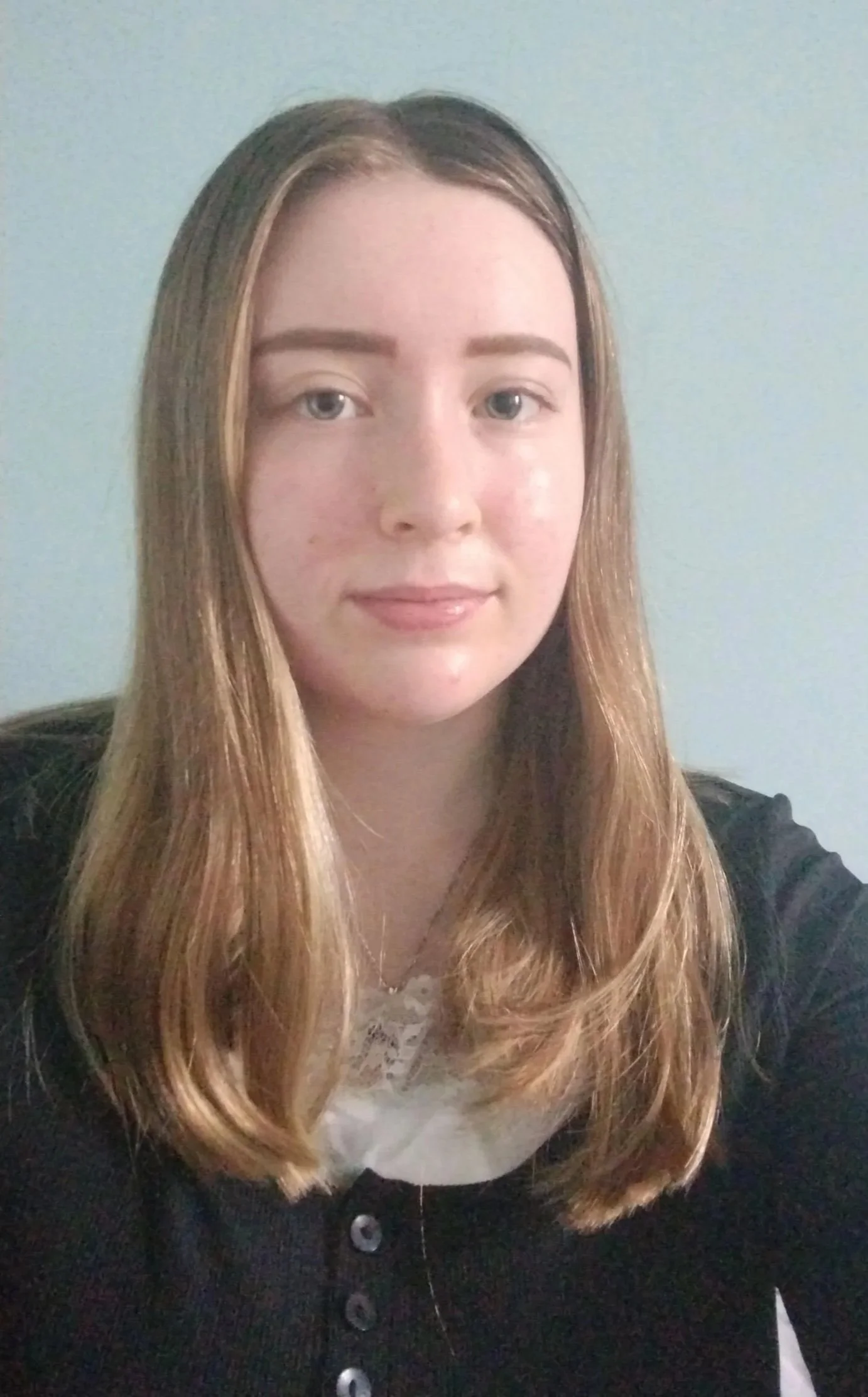 Young woman with long, light brown hair in loose waves, wearing a black cardigan over a white top, against a light gray background.
