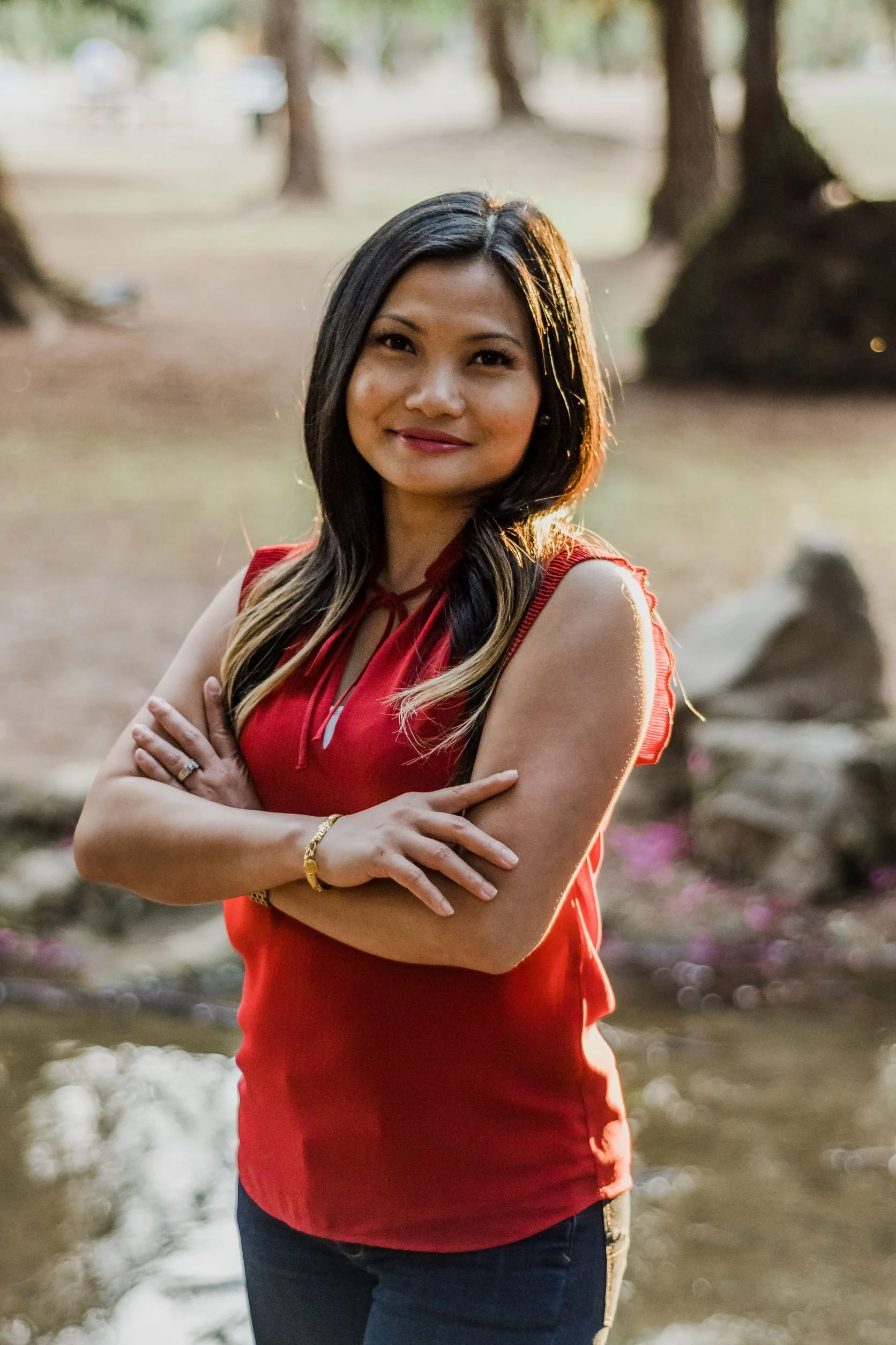 A woman with black and blonde hair wearing a red sleeveless top and dark jeans standing outdoors near a body of water with trees in the background.