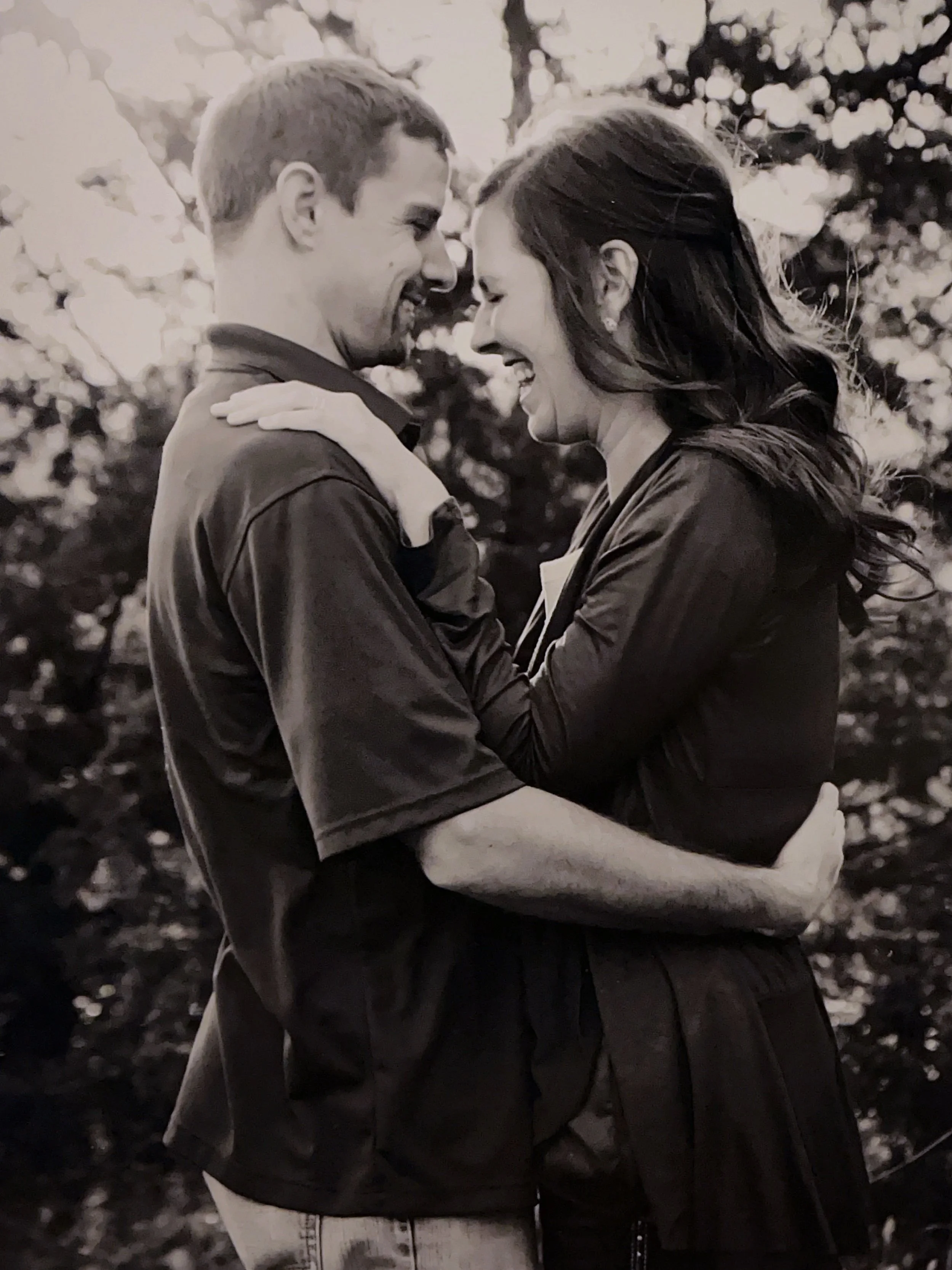 A black and white photo of a couple smiling and touching foreheads with their eyes closed, outdoors with trees in the background.