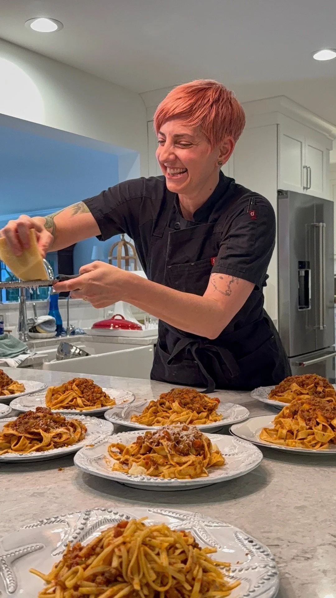 At a Private Chef Lora Plauche private Cooking Class Lora is grating cheese on multiple plates of spaghetti and meat sauce in a modern kitchen.