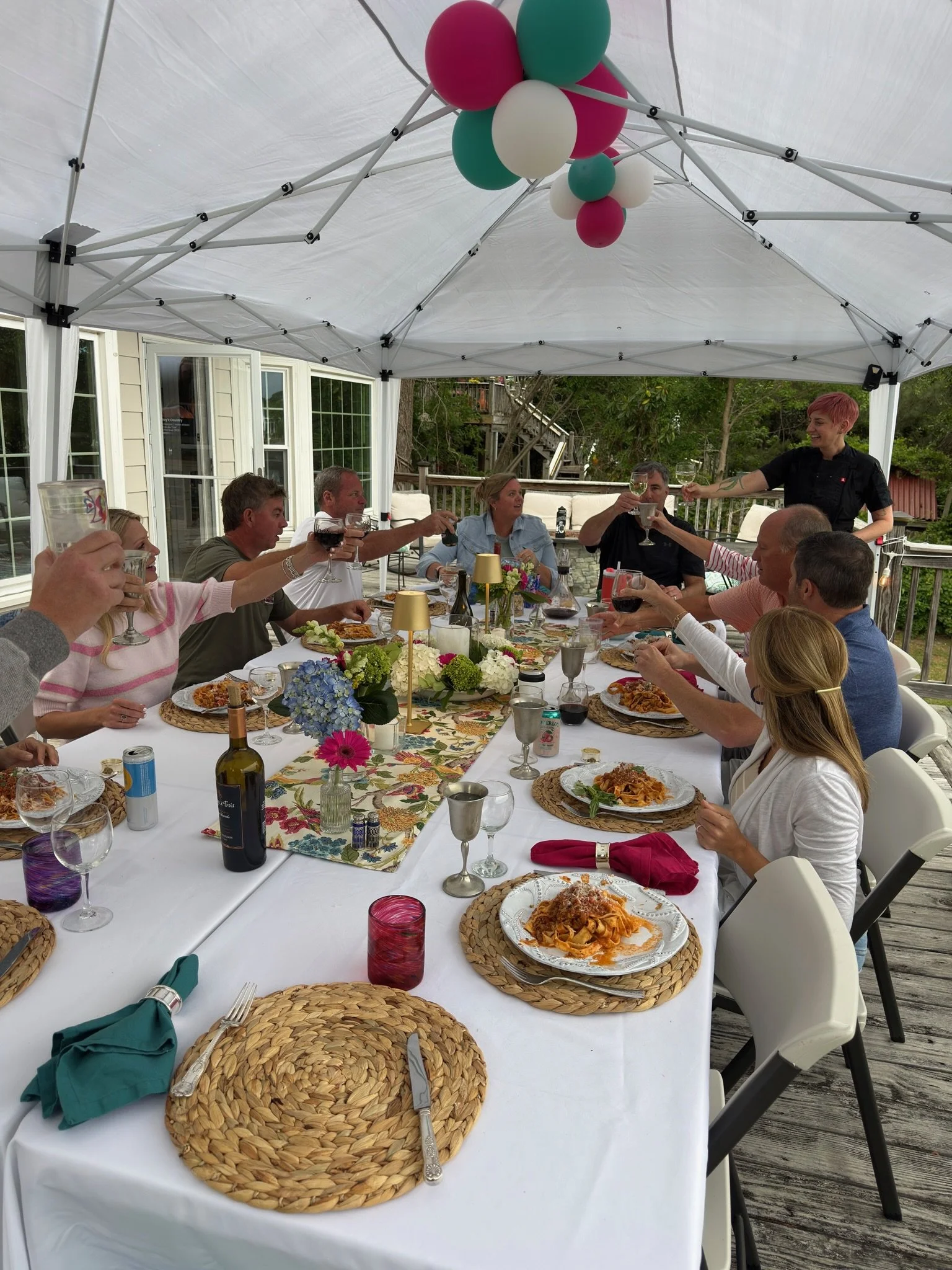 People gathered around a long outdoor dining table under a white canopy at a Private Chef Lora Plauche private event, celebrating with drinks and food, with some pink, white, and green balloons hanging from the canopy.