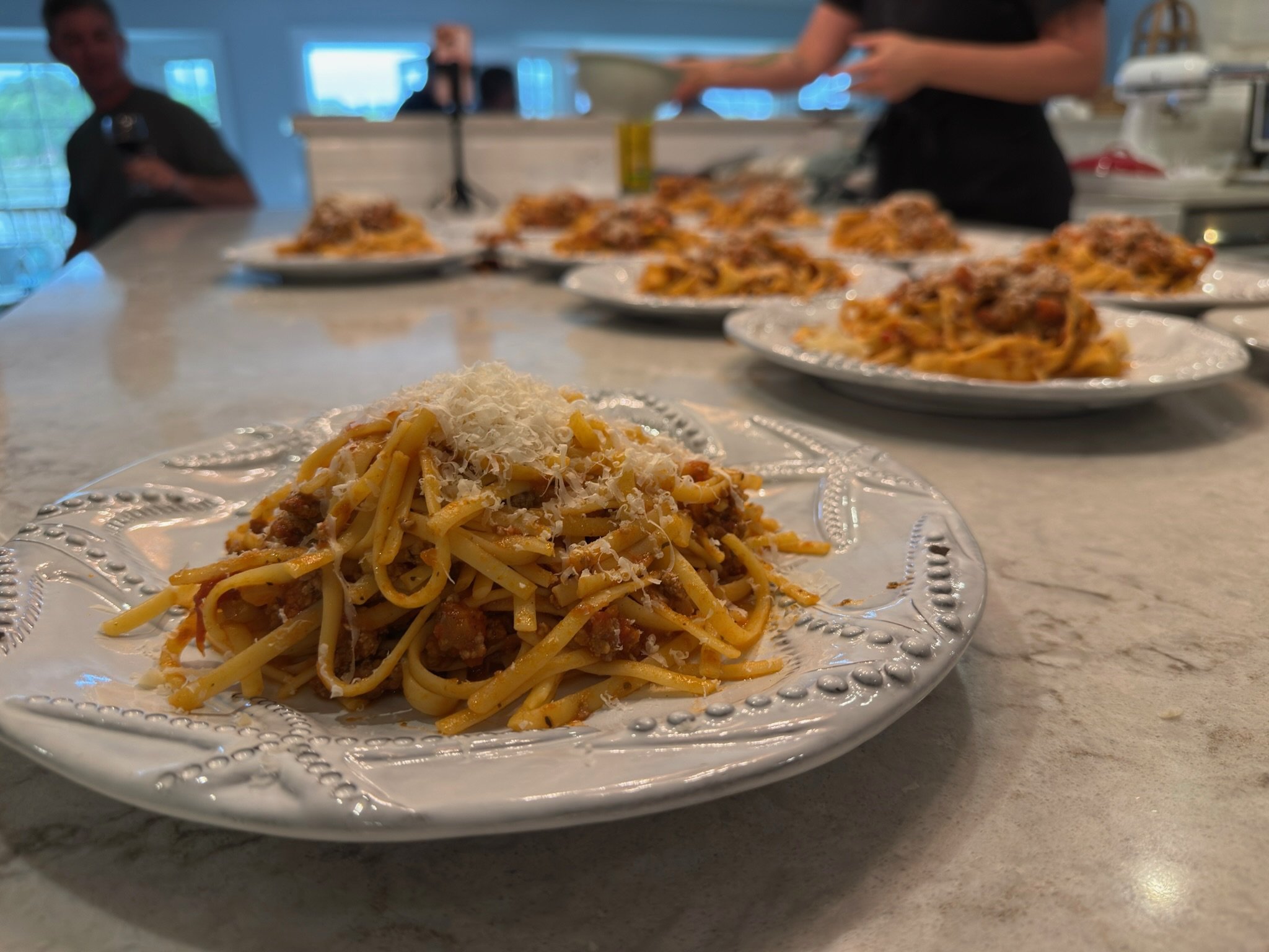 Plate of spaghetti with grated cheese in the foreground, several more plates of spaghetti on a table, people working in the background at a Private Chef Lora Plauche private cooking class event.
